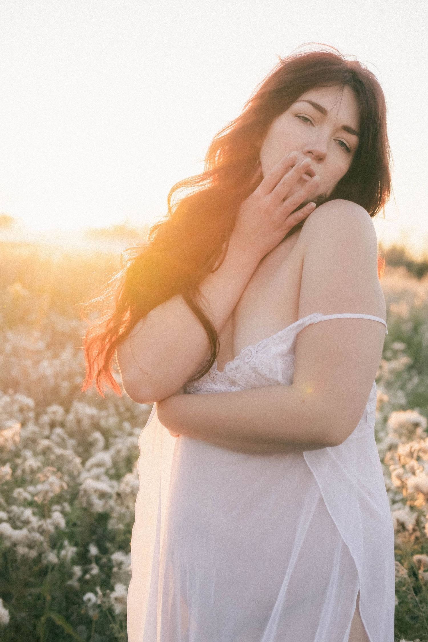 Woman in white lace dress standing in blooming field during golden sunset, long dark hair flowing in warm light.