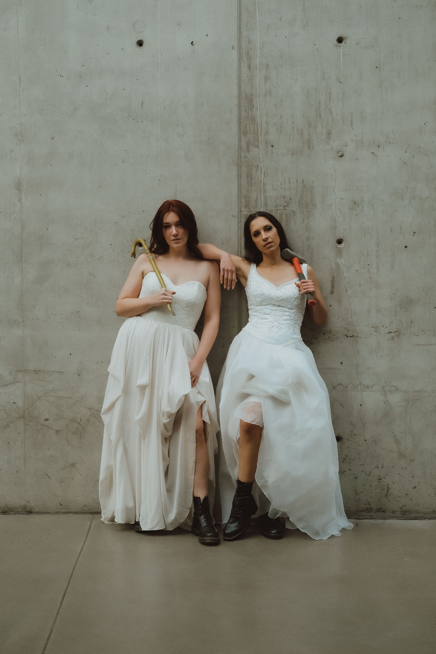 Two women in white wedding dresses and black boots lean against a concrete wall holding tools.