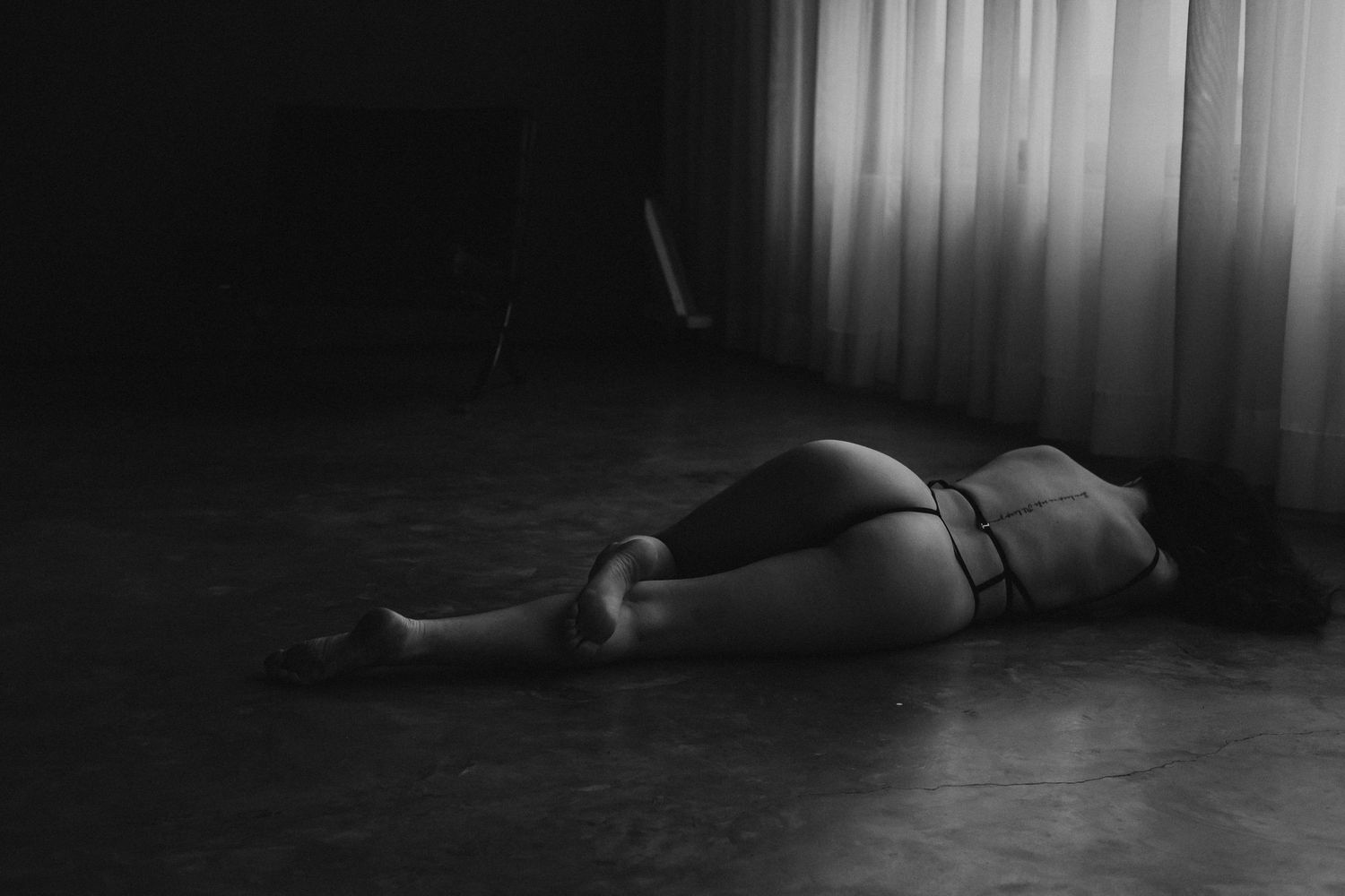 A black and white fine art photograph of a woman lying on a studio floor near sheer curtains in lingerie.