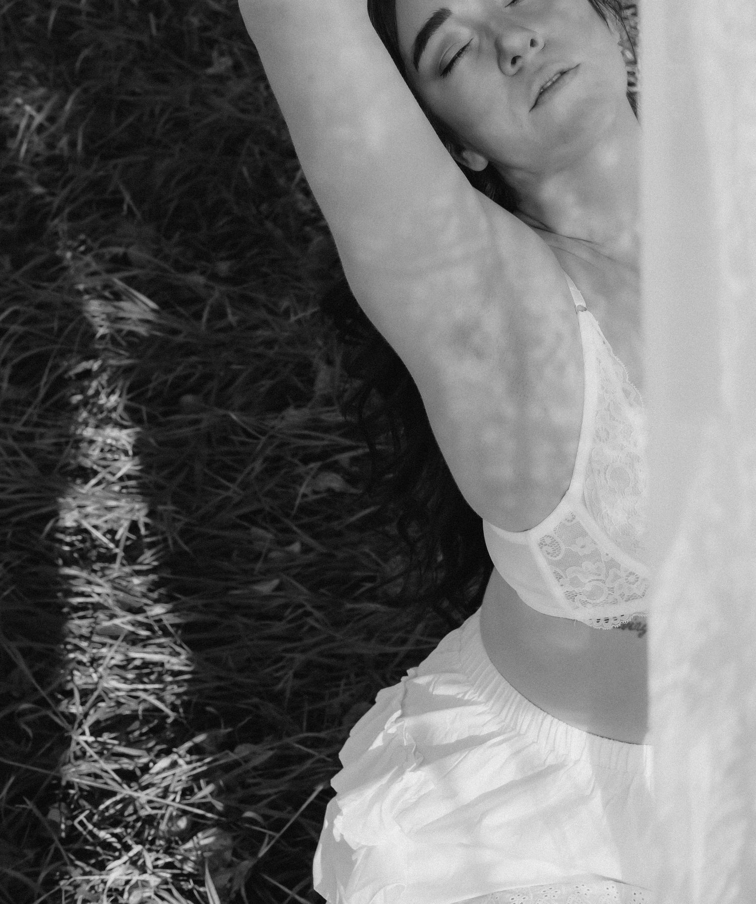 Black and white photo of woman in white lace bra stretching arms in sunlit field.