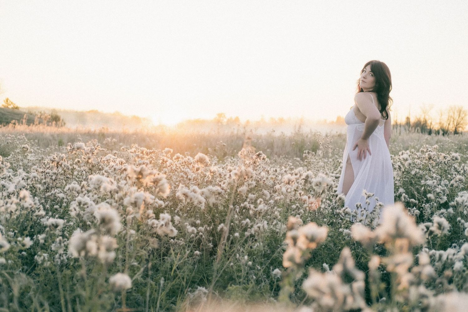 Woman in white dress standing in a field of wildflowers at golden sunset hour.