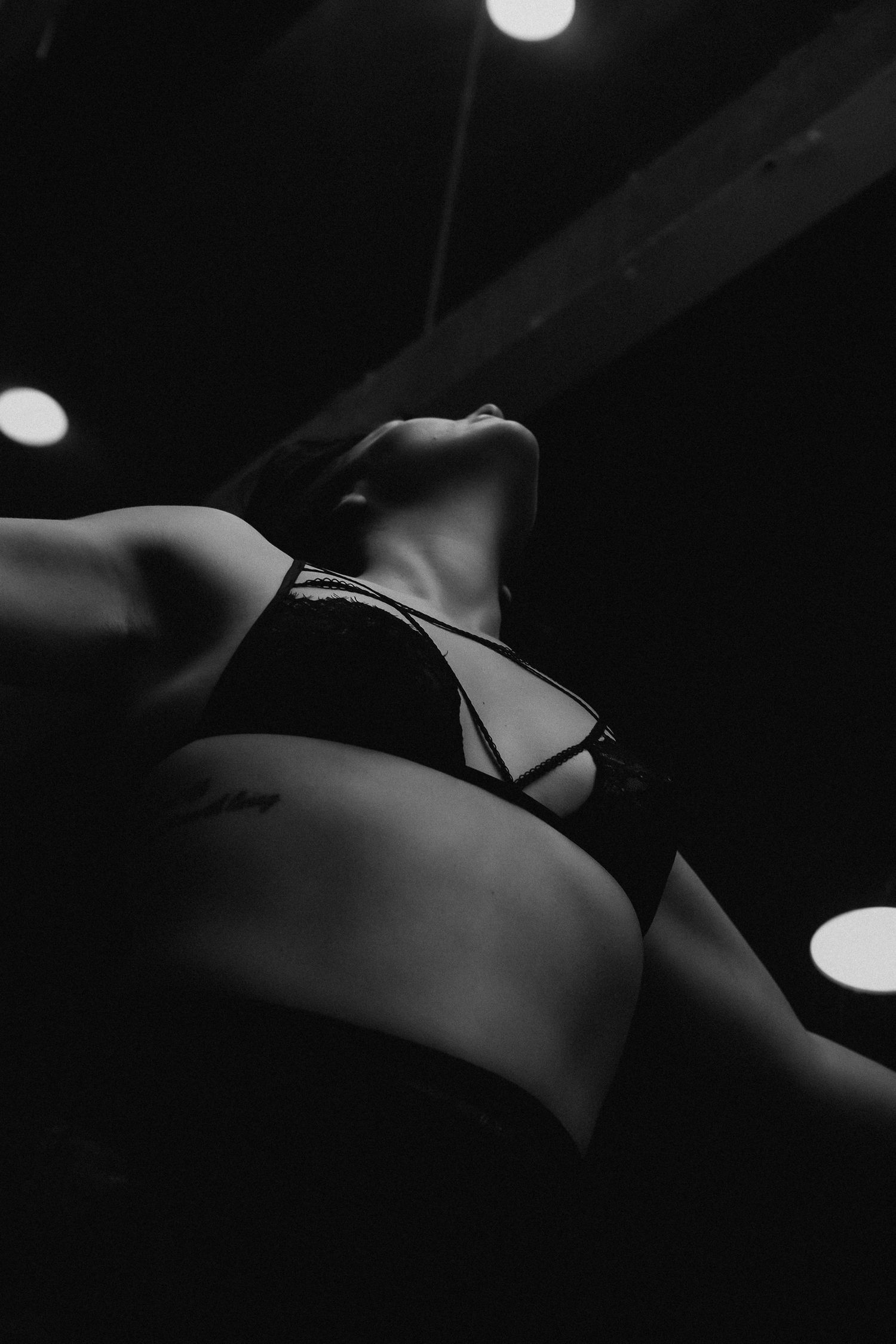 Black and white low-angle photo of a woman in a black bikini top, with dramatic ceiling lights in the background.