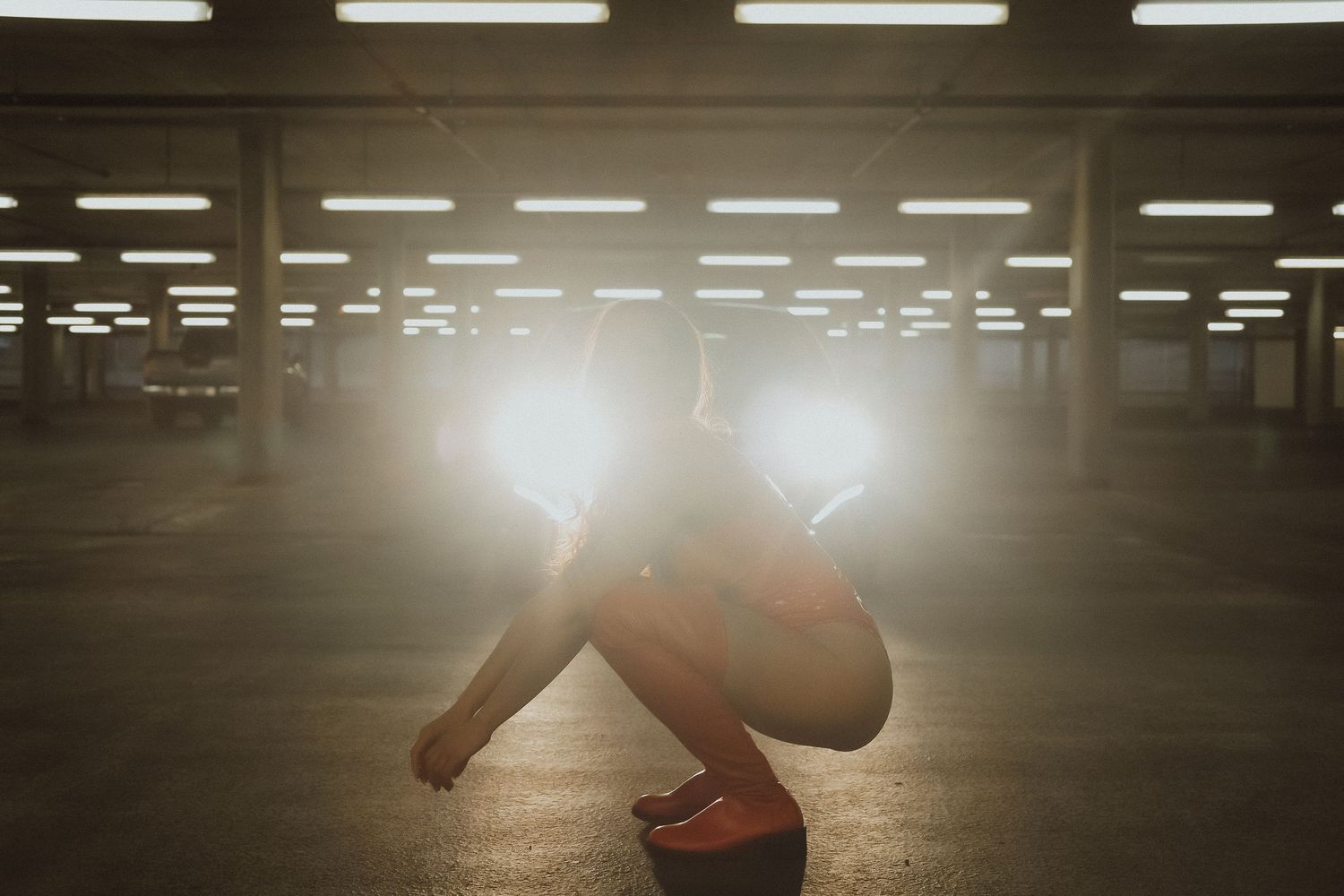 A woman crouching in an empty parking garage with dramatic backlighting creating a glowing silhouette effect.
