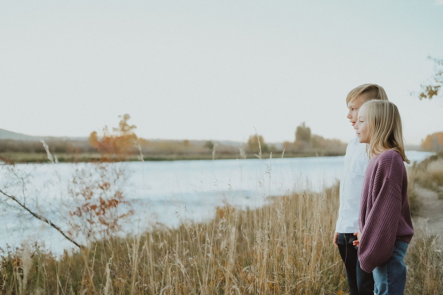 Two girls standing near a lake surrounded by tall golden grass during autumn.