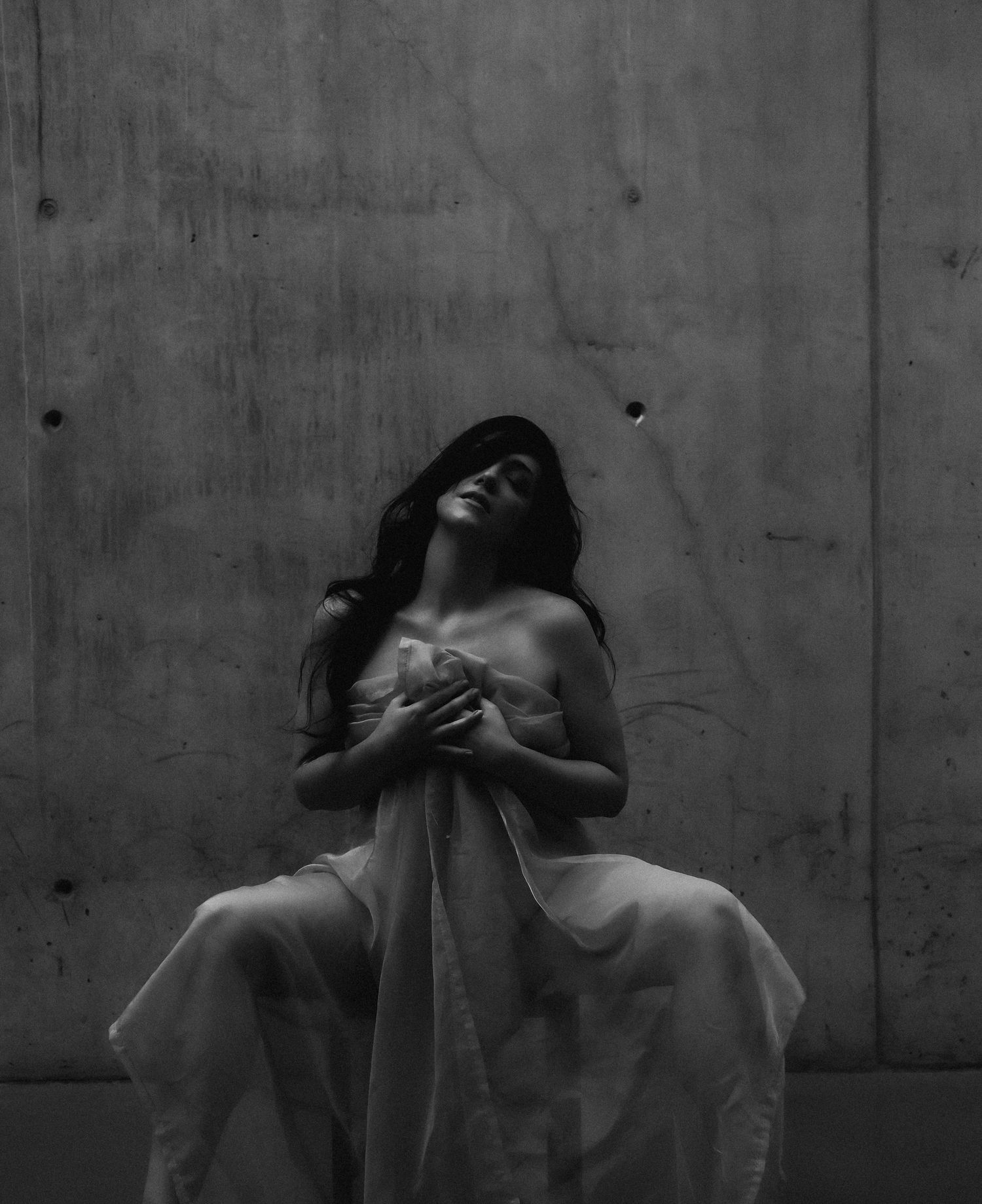 Black and white photo of a woman in a flowing white dress, head tilted back, hands crossed over chest, against a concrete wall.