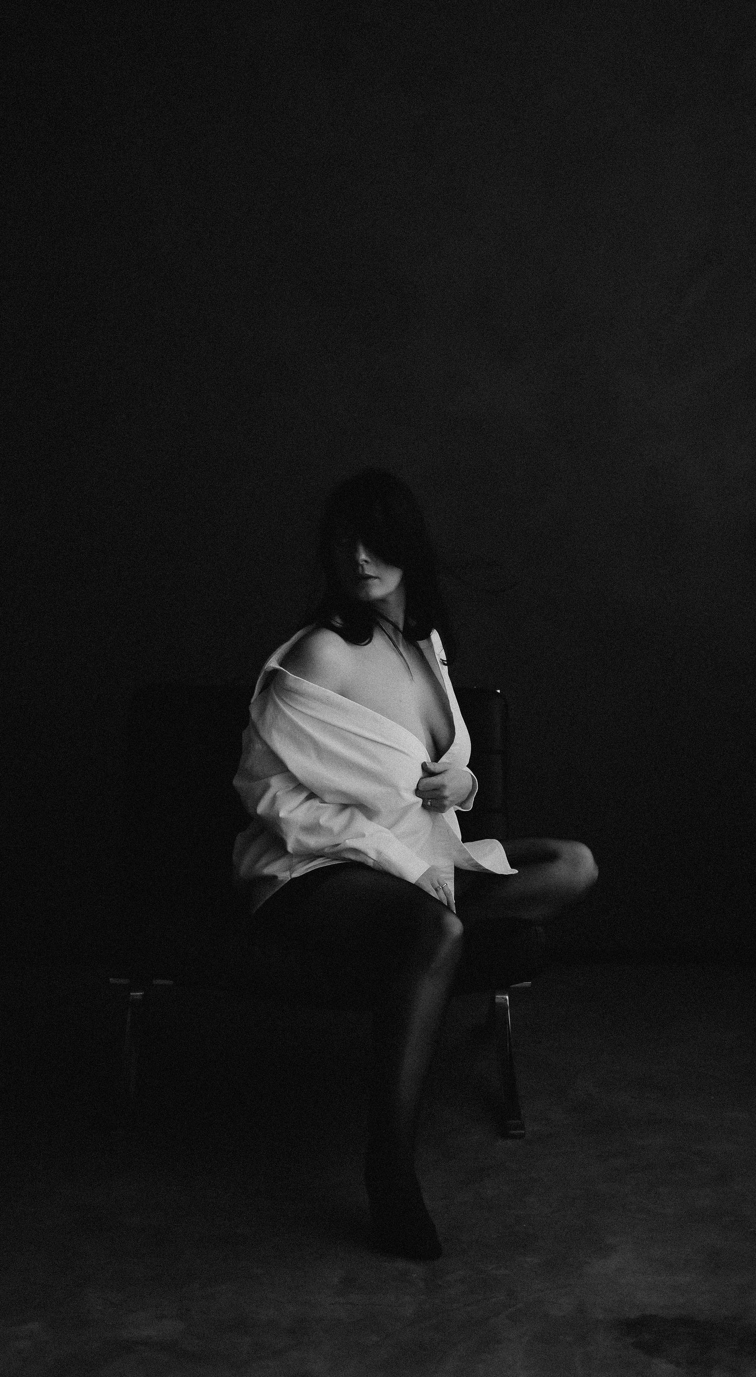 Moody black and white photo of a woman in an oversized white shirt seated in a dark studio.