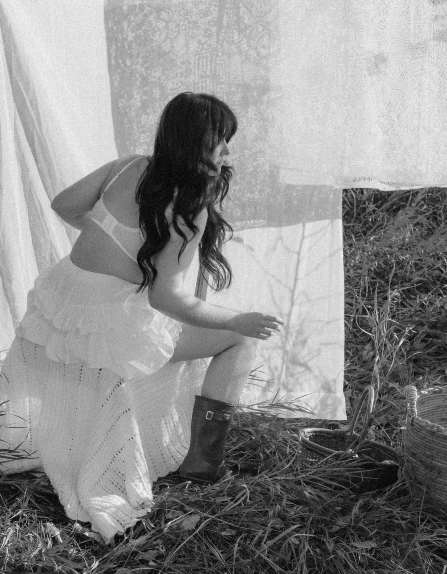 Black and white photo of a woman in a white dress and boots crouching near hanging white linens outdoors.