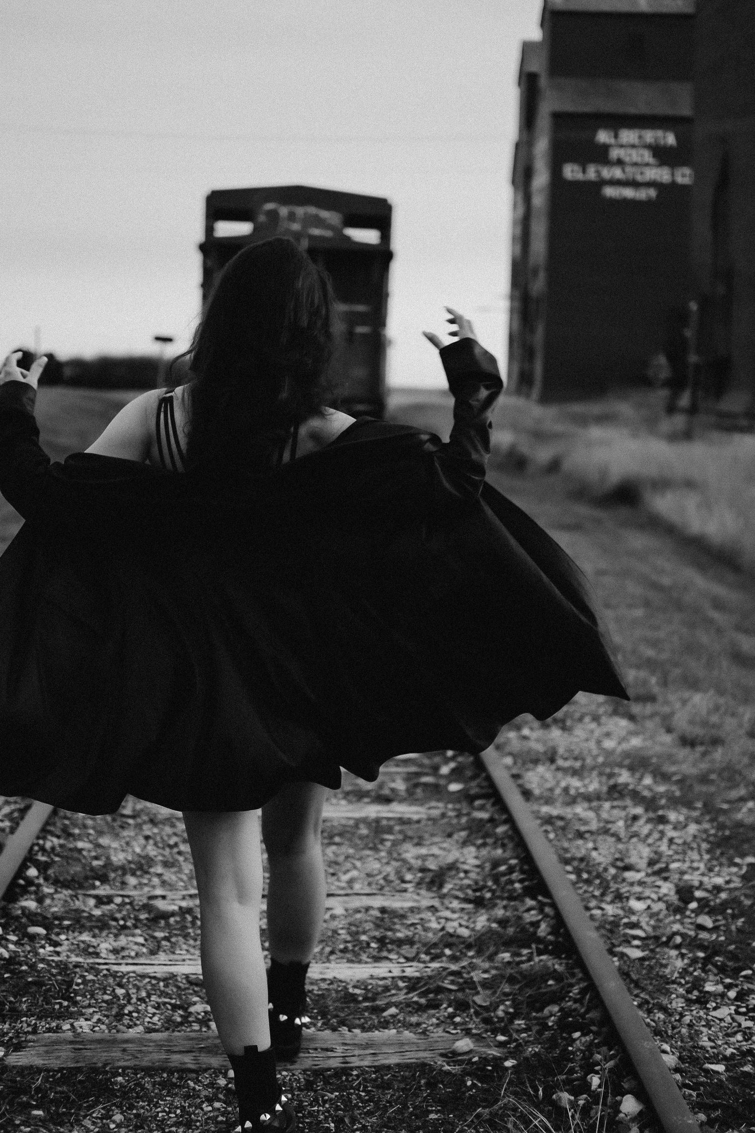 Black and white photo of a woman in a black dress walking on railroad tracks near Alberta Pool Elevators.