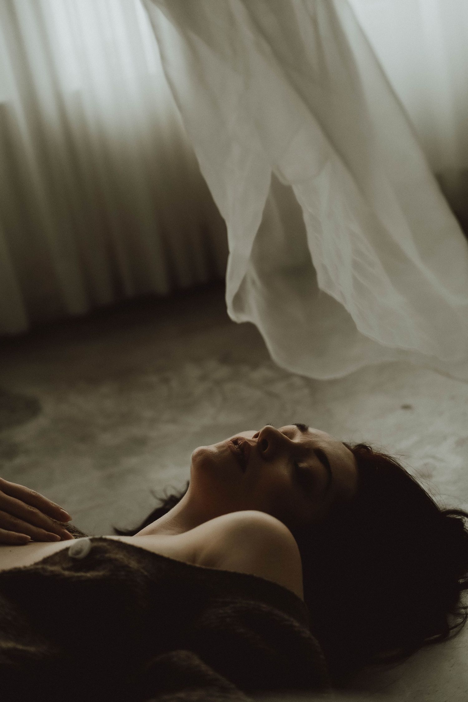A young woman with dark hair lies peacefully on the floor with eyes closed, white fabric floating above her in soft light.