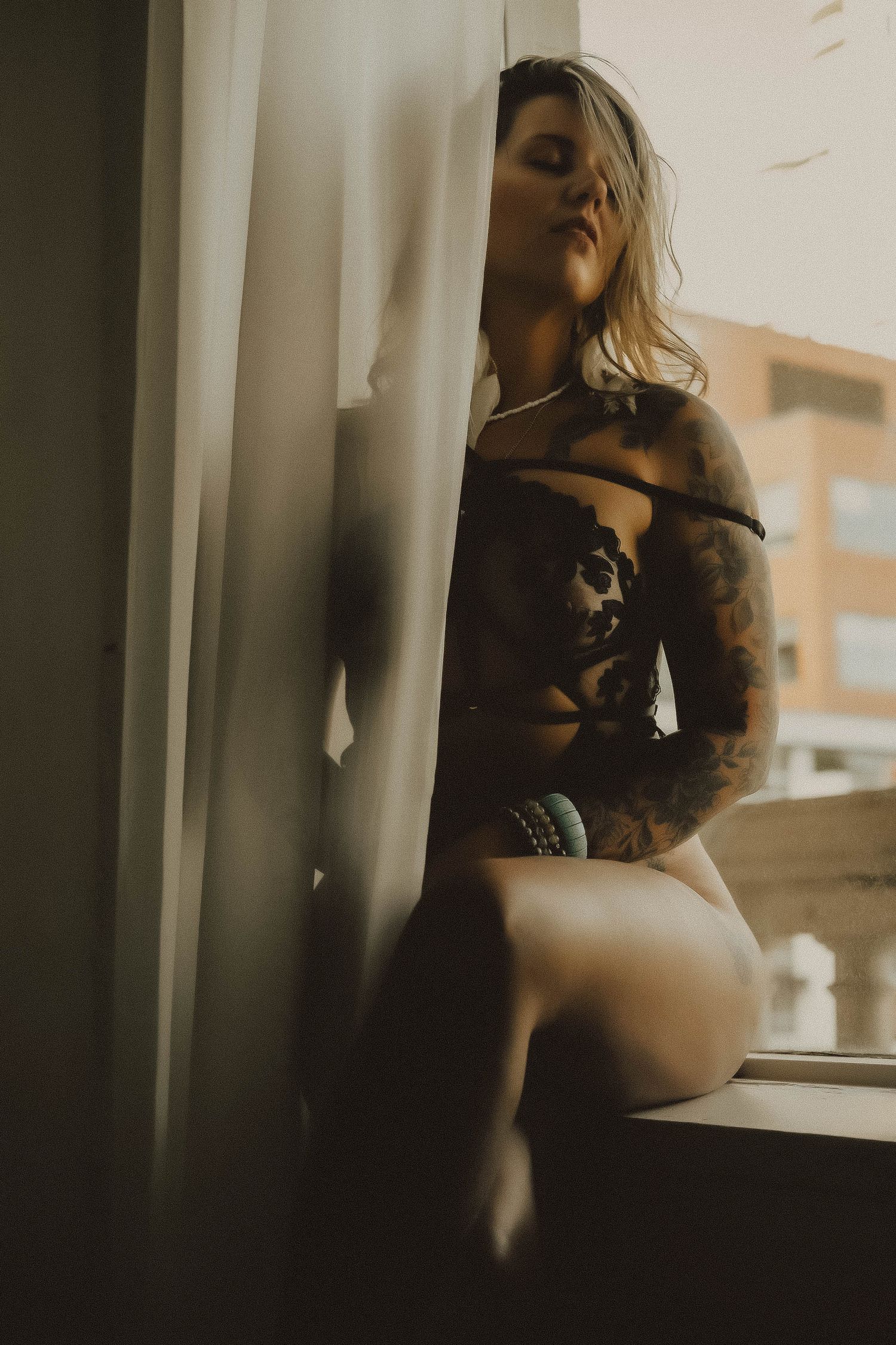 Tattooed blonde woman in dark lingerie posing by a sunlit window with sheer curtains in moody boudoir photo.