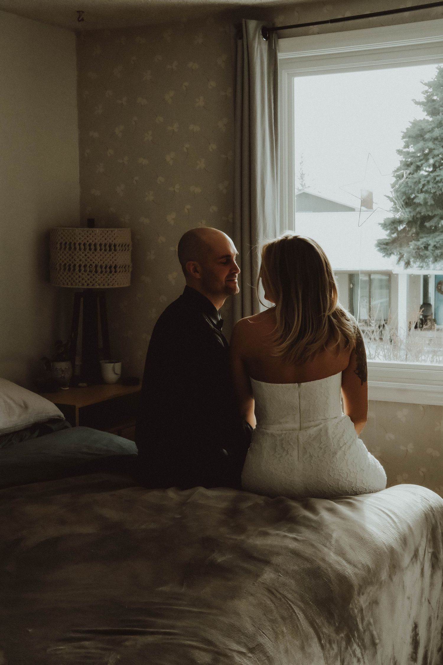 Bride and groom sitting on a bed together near a window in a softly lit bedroom.