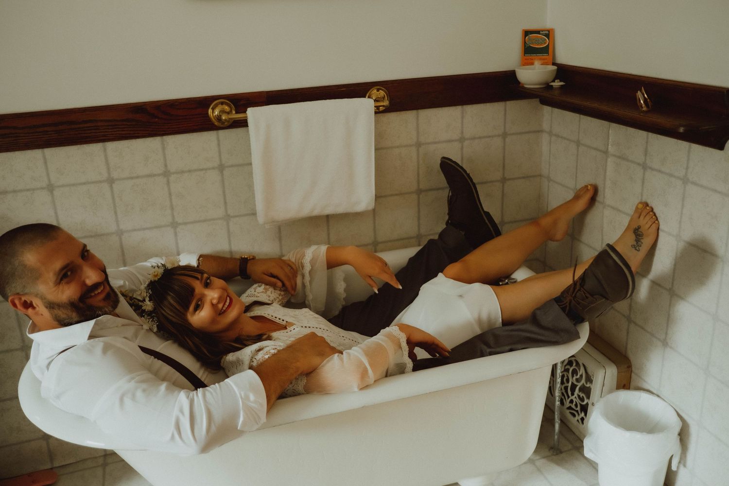 Couple in wedding attire laughing together while relaxing in a vintage bathtub in a tiled bathroom.