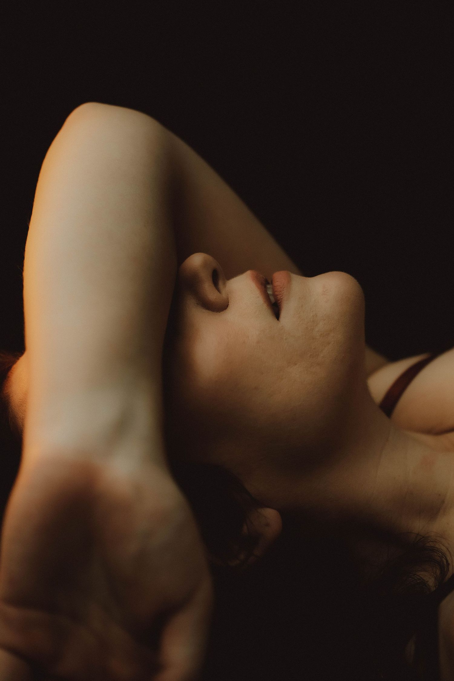 A woman reclines with her arm raised, head tilted back, in dramatic low-key lighting against a dark background.