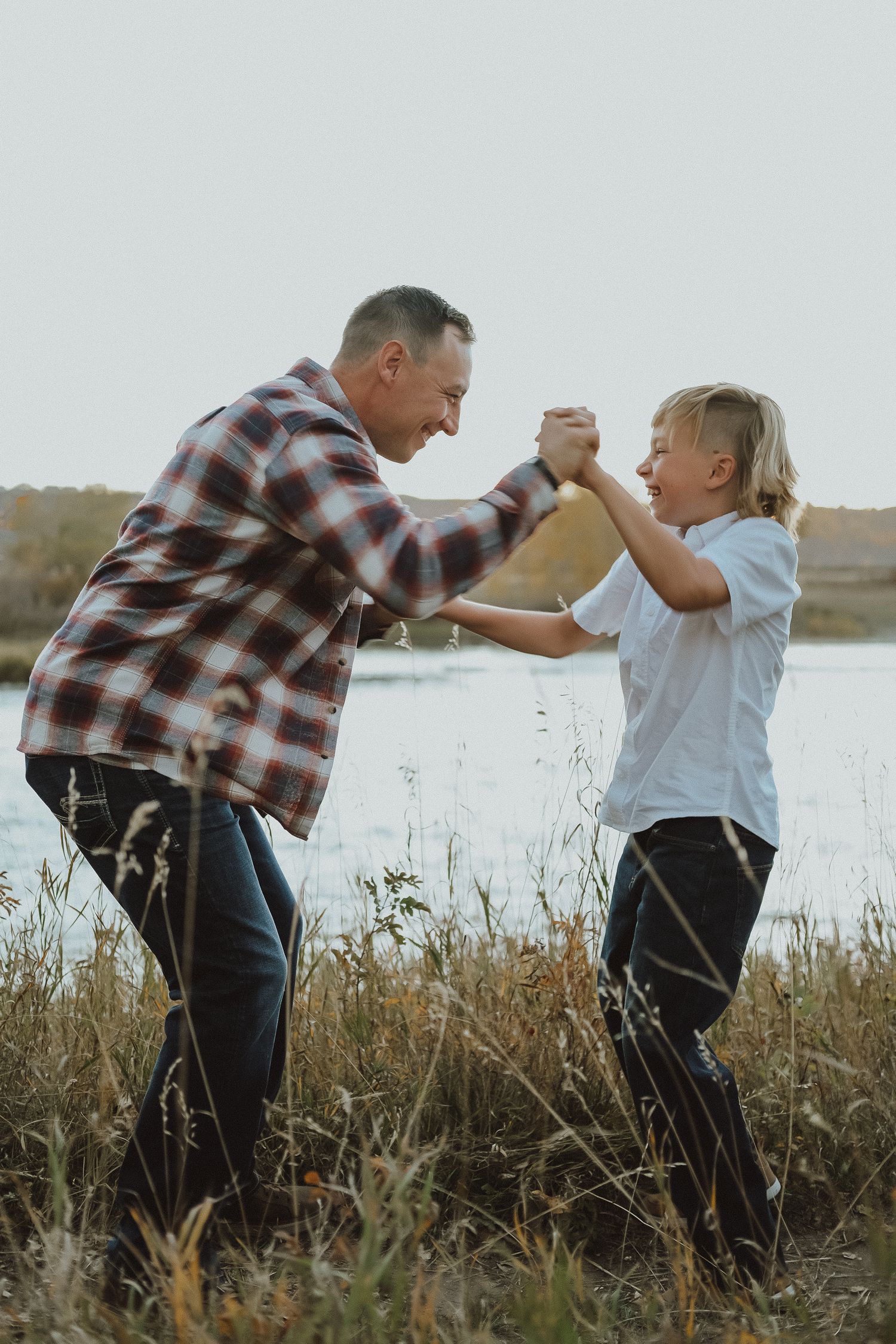 Father and son holding hands and playing together in a grassy field near a lake on an autumn day.
