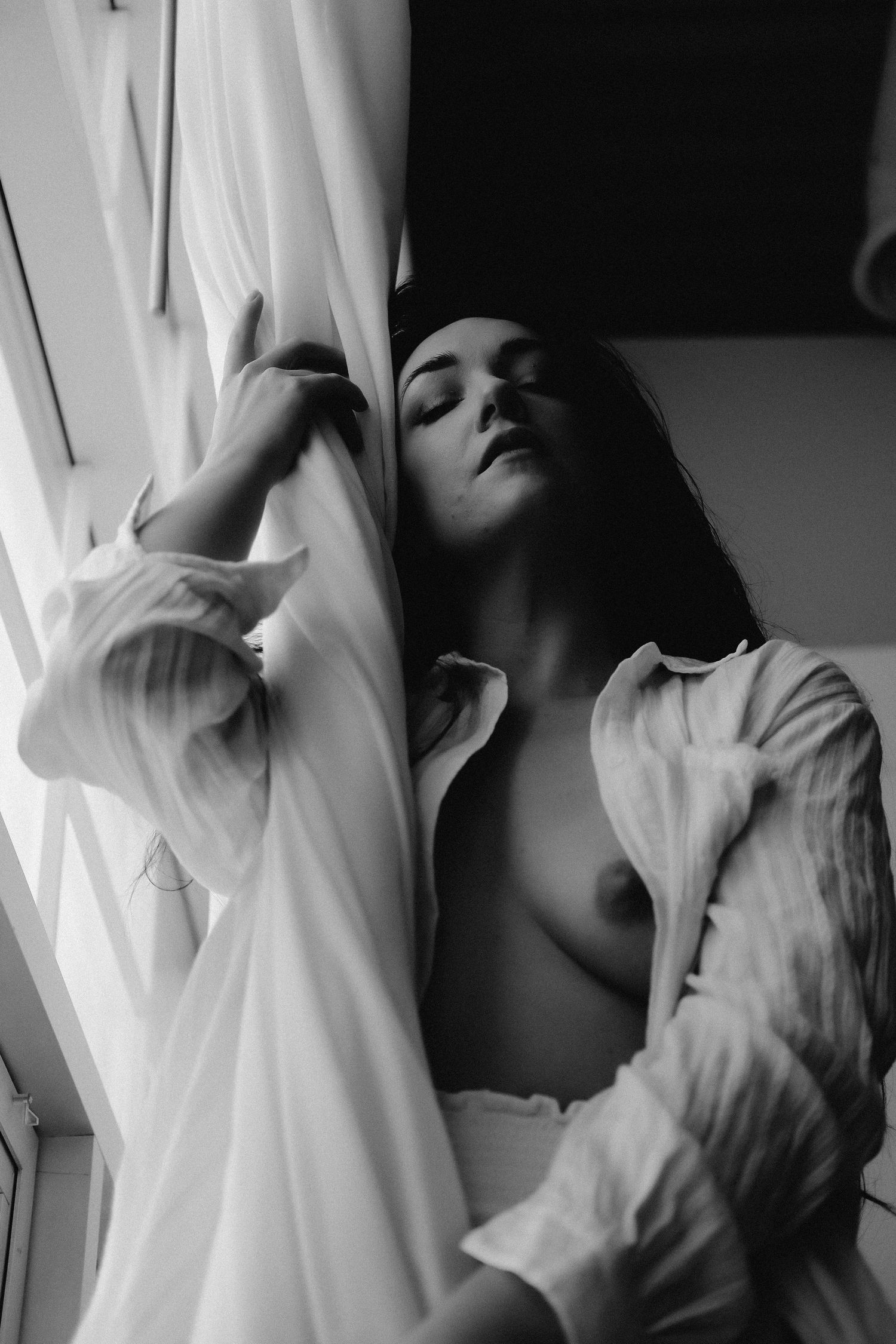 Black and white artistic boudoir photo of a woman in an open white shirt leaning against sheer curtains by a window.