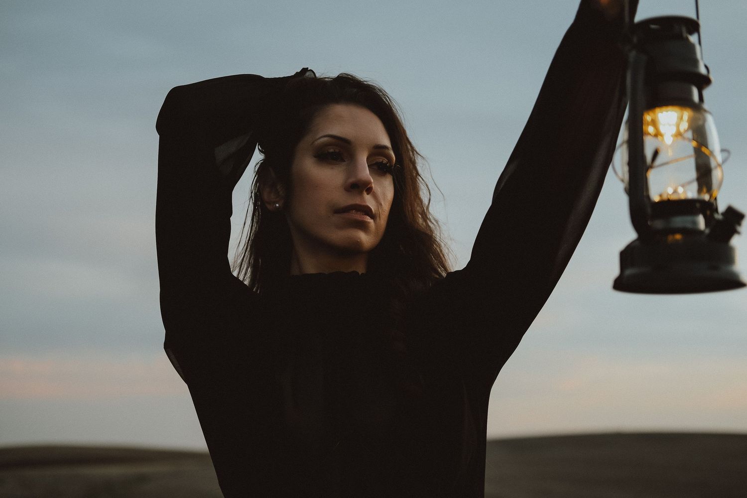 Woman in black outfit holds vintage lantern aloft against dusky sky in dramatic outdoor portrait.