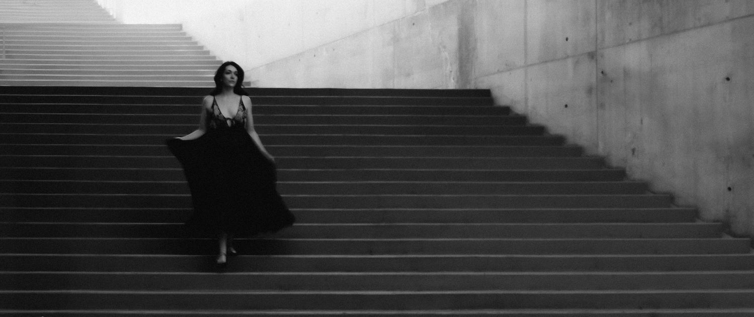 A woman in a flowing black gown descends grand concrete steps in a dramatic black and white fashion photograph.