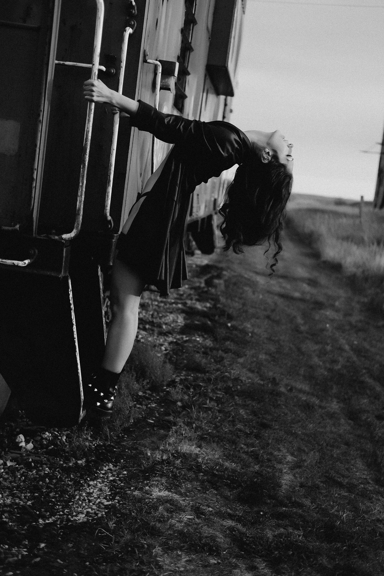 Black and white photo of a woman in a black dress leaning out from a moving train door, hair flowing in the wind.