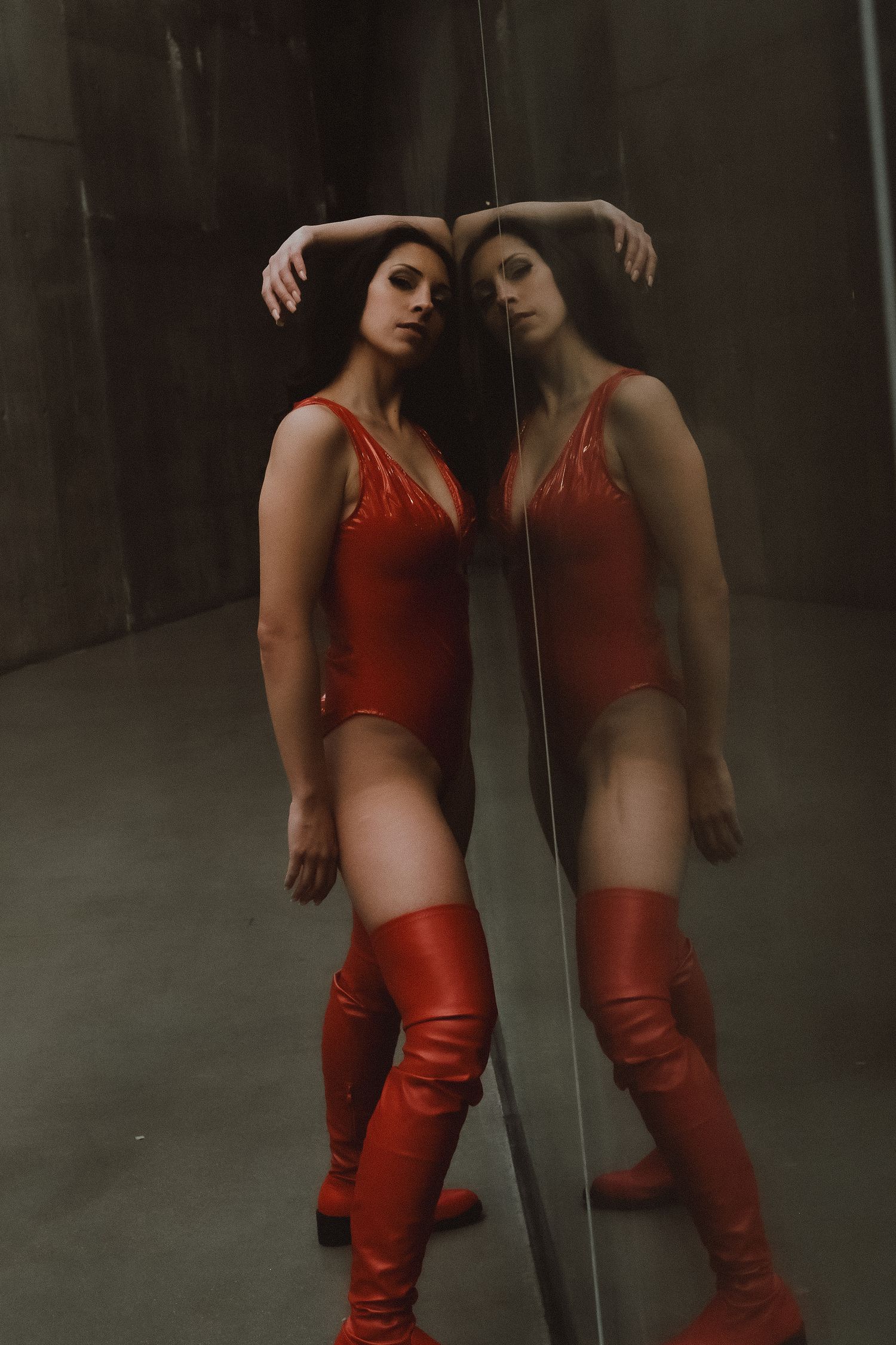 Woman in red bodysuit and thigh-high boots poses against a mirror, creating a striking reflection in a dark room.