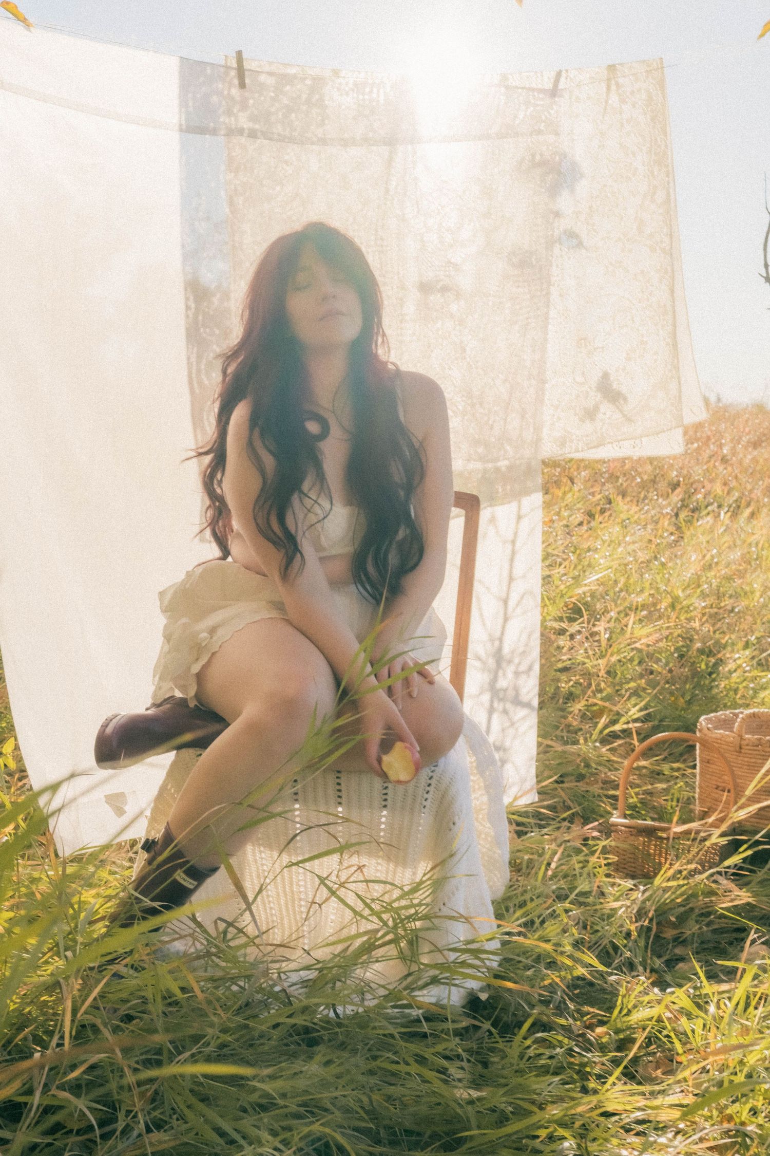Woman in white dress sitting on chair in sunlit field with white linens hanging on clothesline behind her.