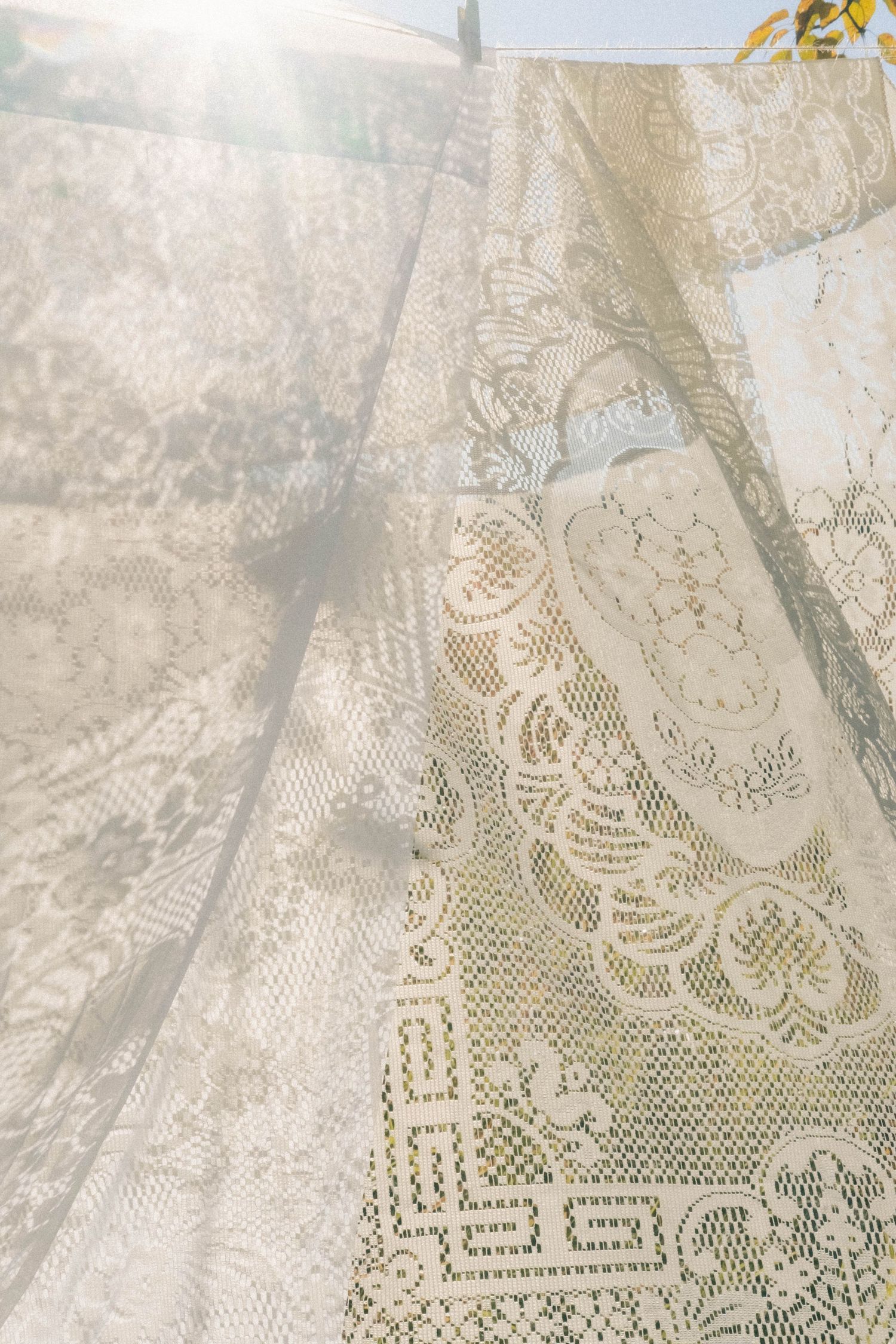 Sunlit vintage cream lace curtains hanging outdoors with intricate floral and geometric patterns.