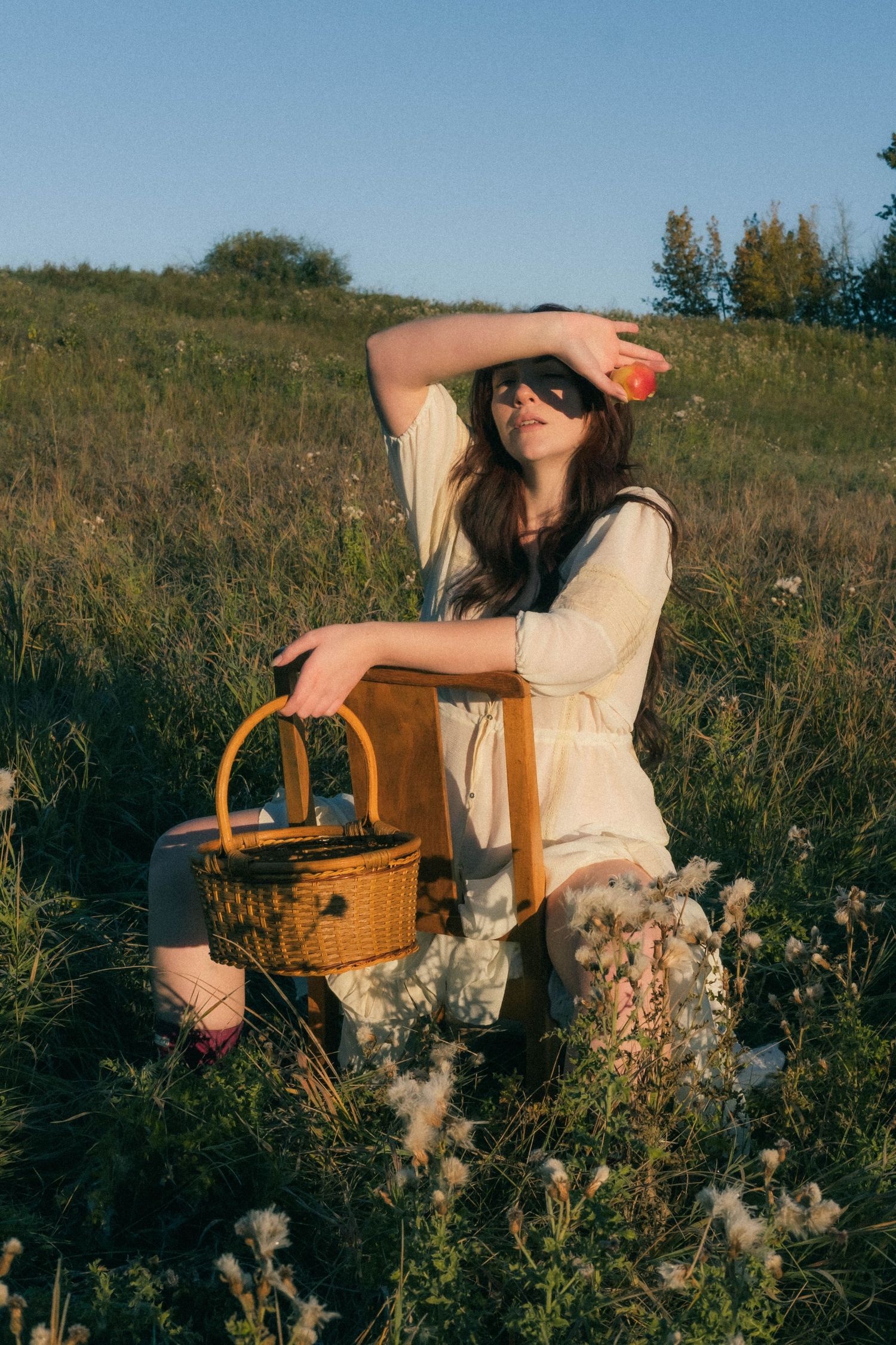 Woman in cream dress sitting in a sunny meadow, shielding her eyes while holding a wicker picnic basket.