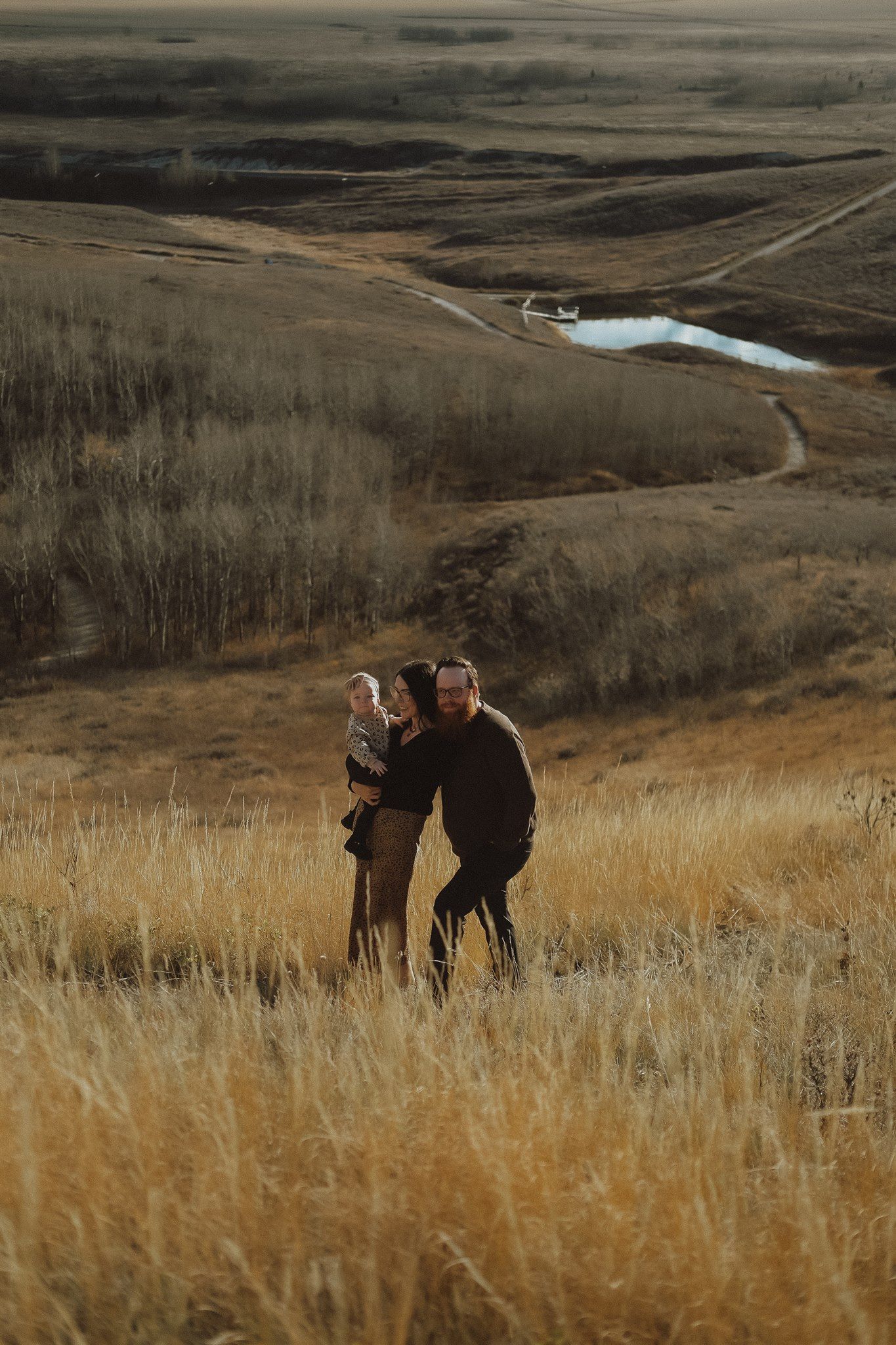 Two people holding a child in golden prairie grass with a winding river valley landscape in the background.