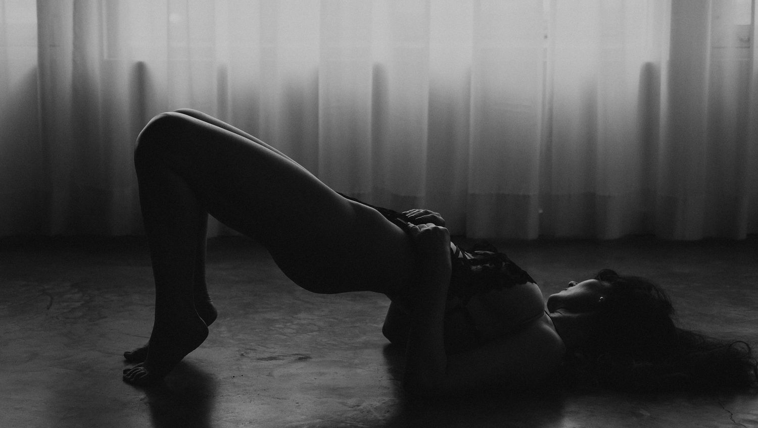 Black and white silhouette of woman in bridge pose on floor before sheer white curtains.