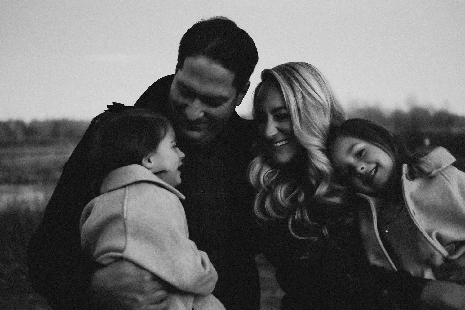 Black and white photo of a loving family of four laughing together outdoors in natural setting.