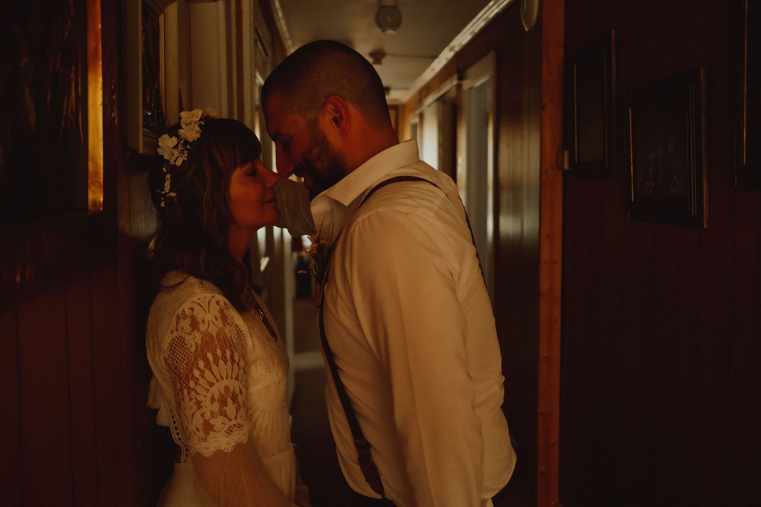A couple shares an intimate moment in a warmly lit hallway, the bride wearing a floral crown and lace dress.