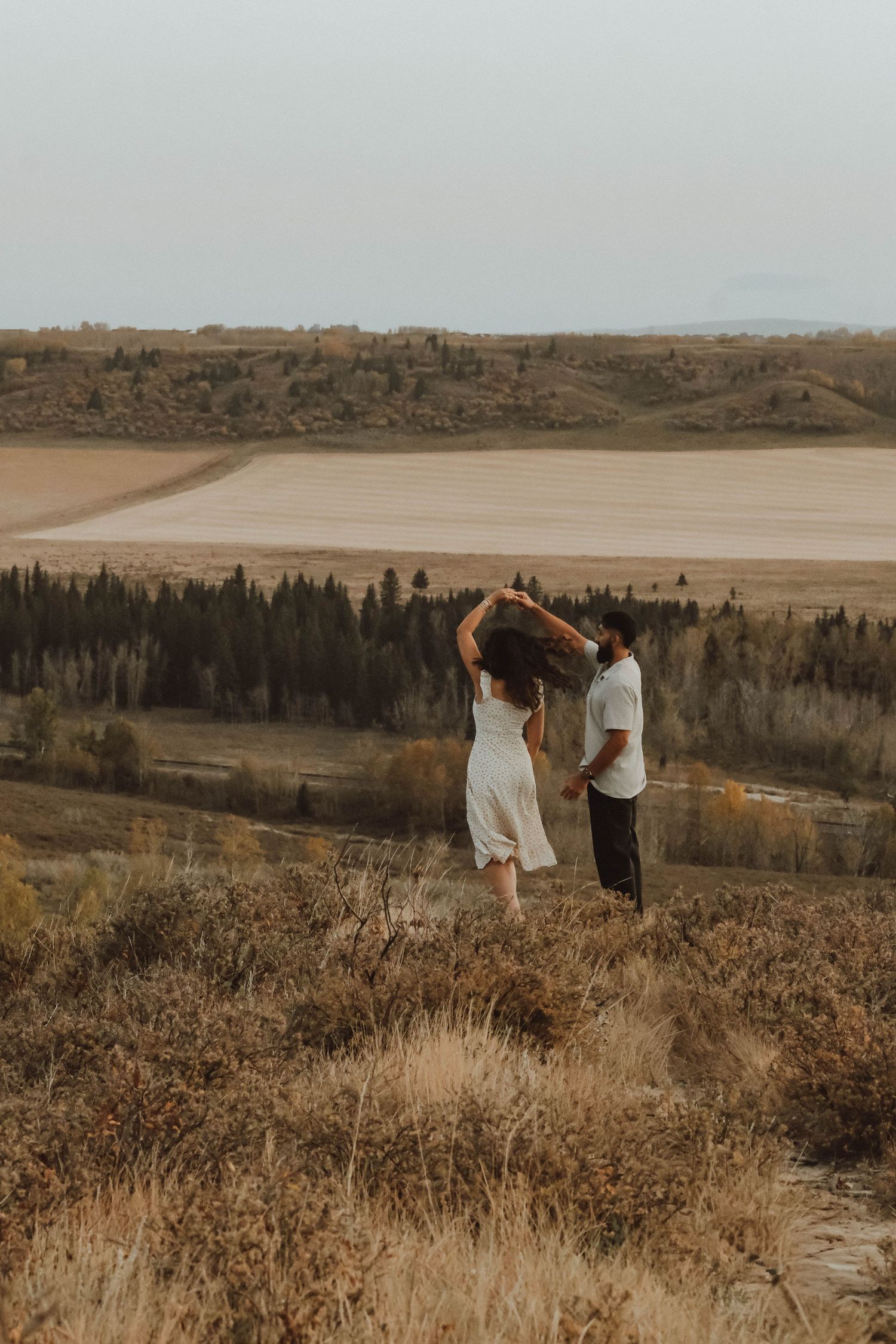 Couple dancing on a hilltop overlooking vast farmland and evergreen trees in a moody outdoor setting.
