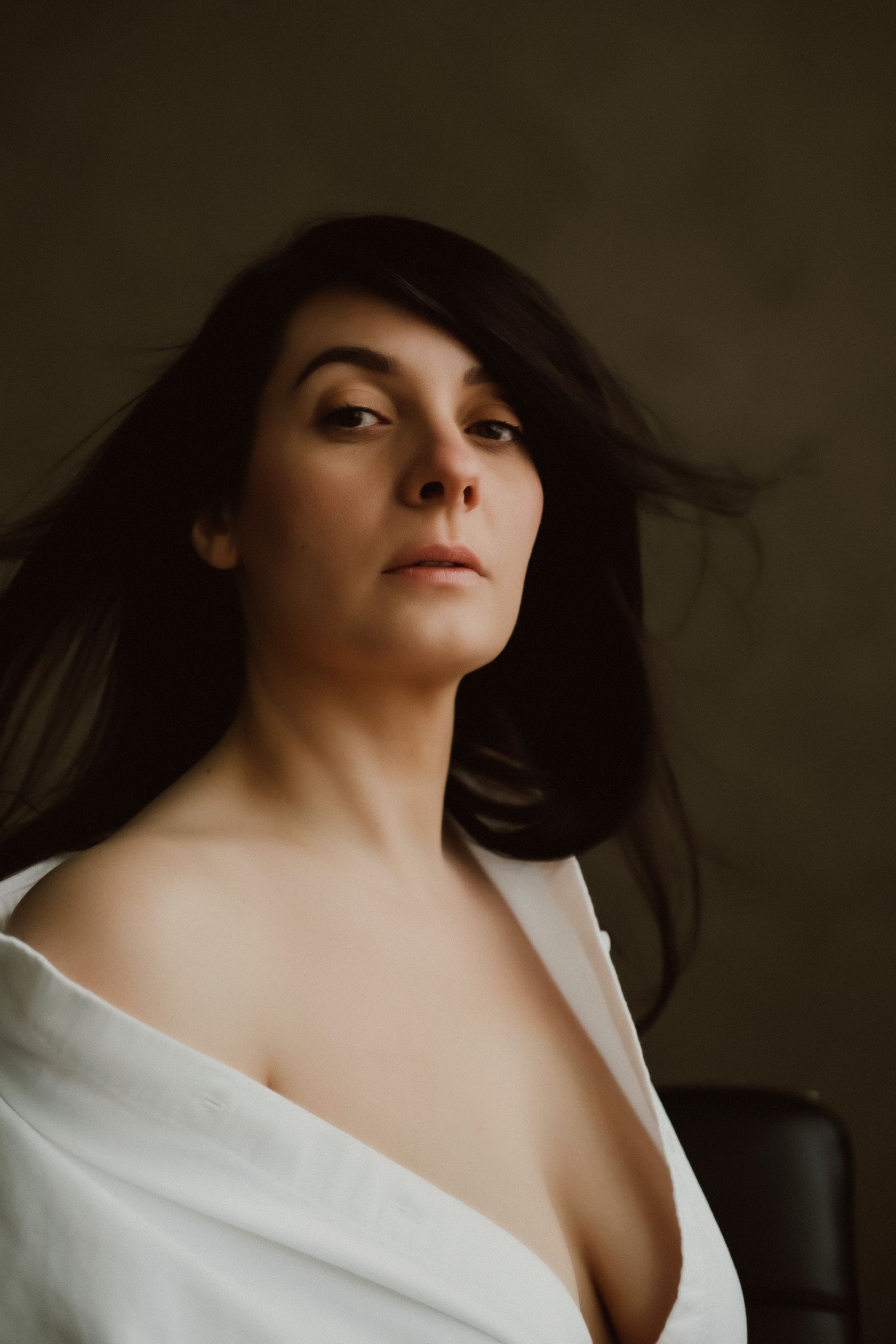 Close-up portrait of dark-haired woman in white shirt with wind-blown hair on dark background.