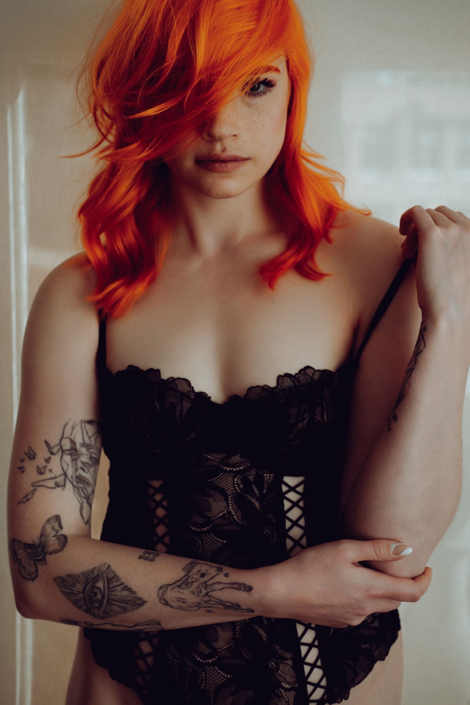 Young woman with vibrant orange hair and tattoos wearing black lace lingerie posing indoors.