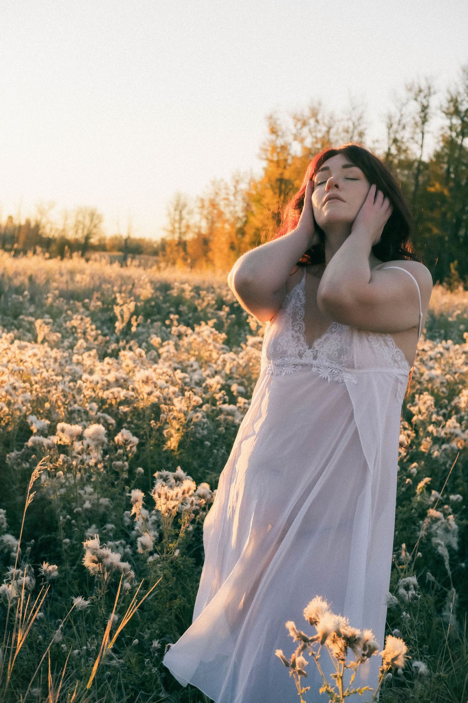 Woman in white dress standing in golden field at sunset, hands framing face, eyes closed, trees in background.