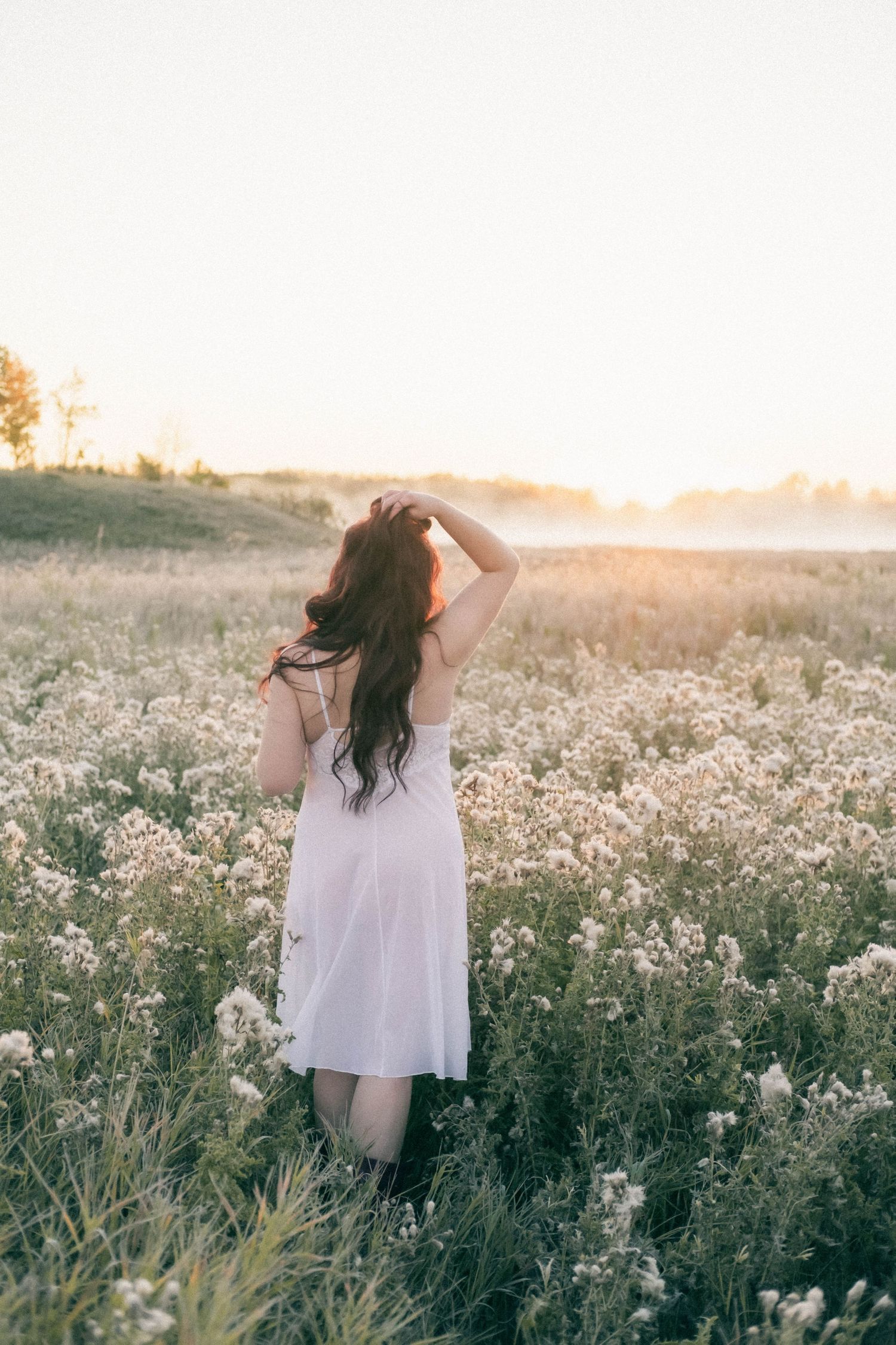Woman in white sundress kneeling in misty wildflower field during golden sunrise.