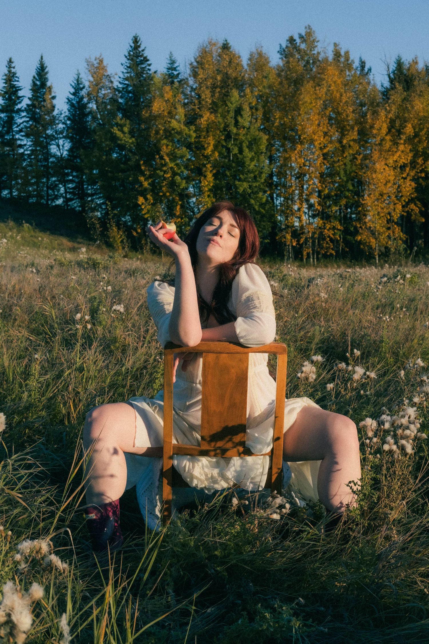 Woman sitting on chair in field holding apple with autumn trees behind.