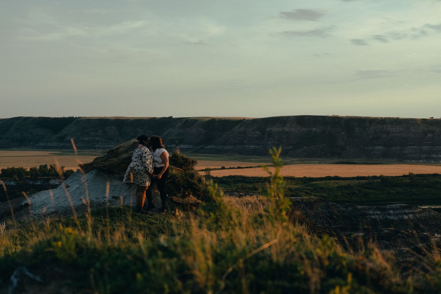 Two people sitting close together on a hillside overlooking a breathtaking golden prairie landscape.