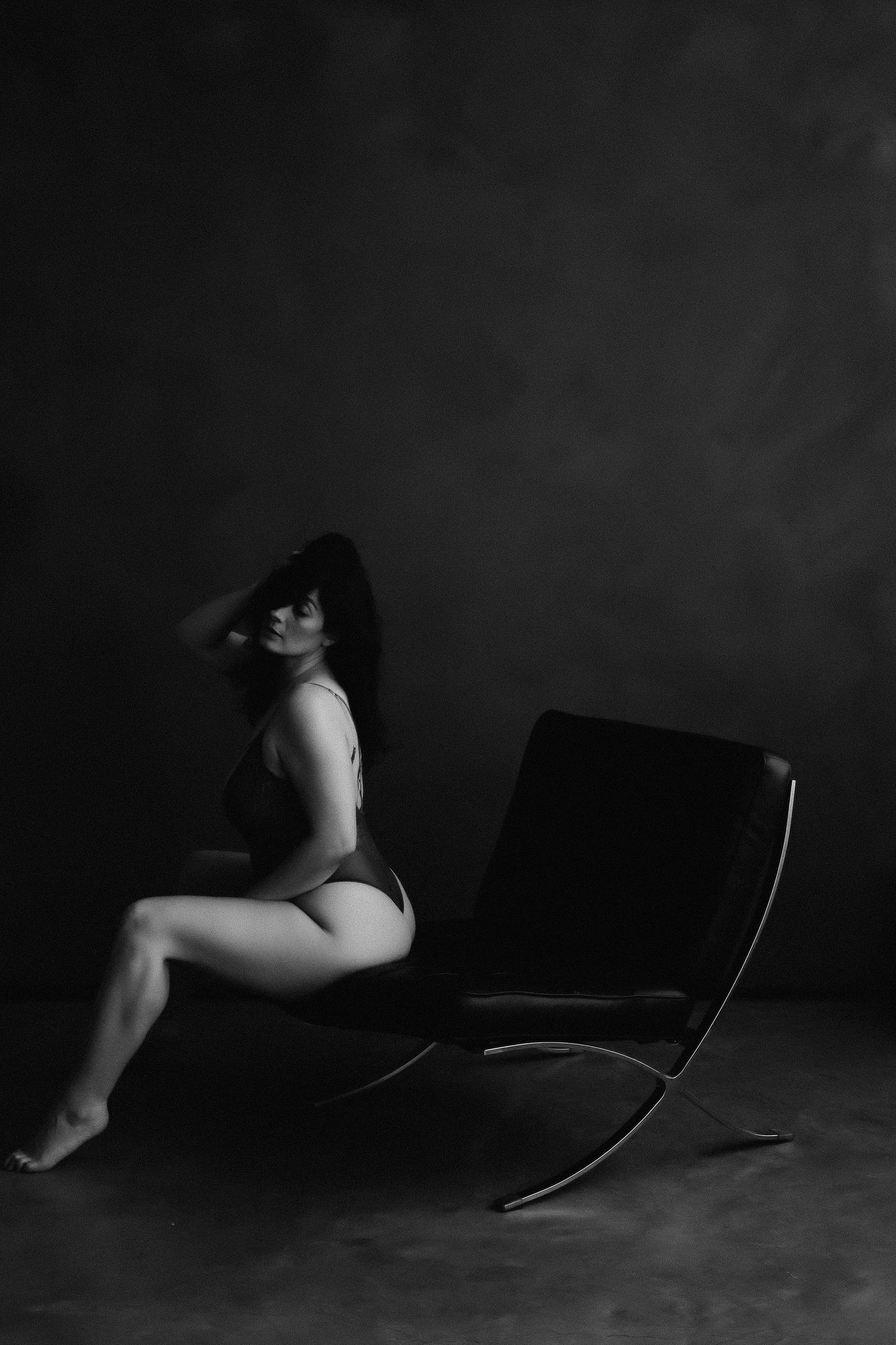 Black and white artistic photo of woman posing in lingerie beside modern leather chair in dark studio.