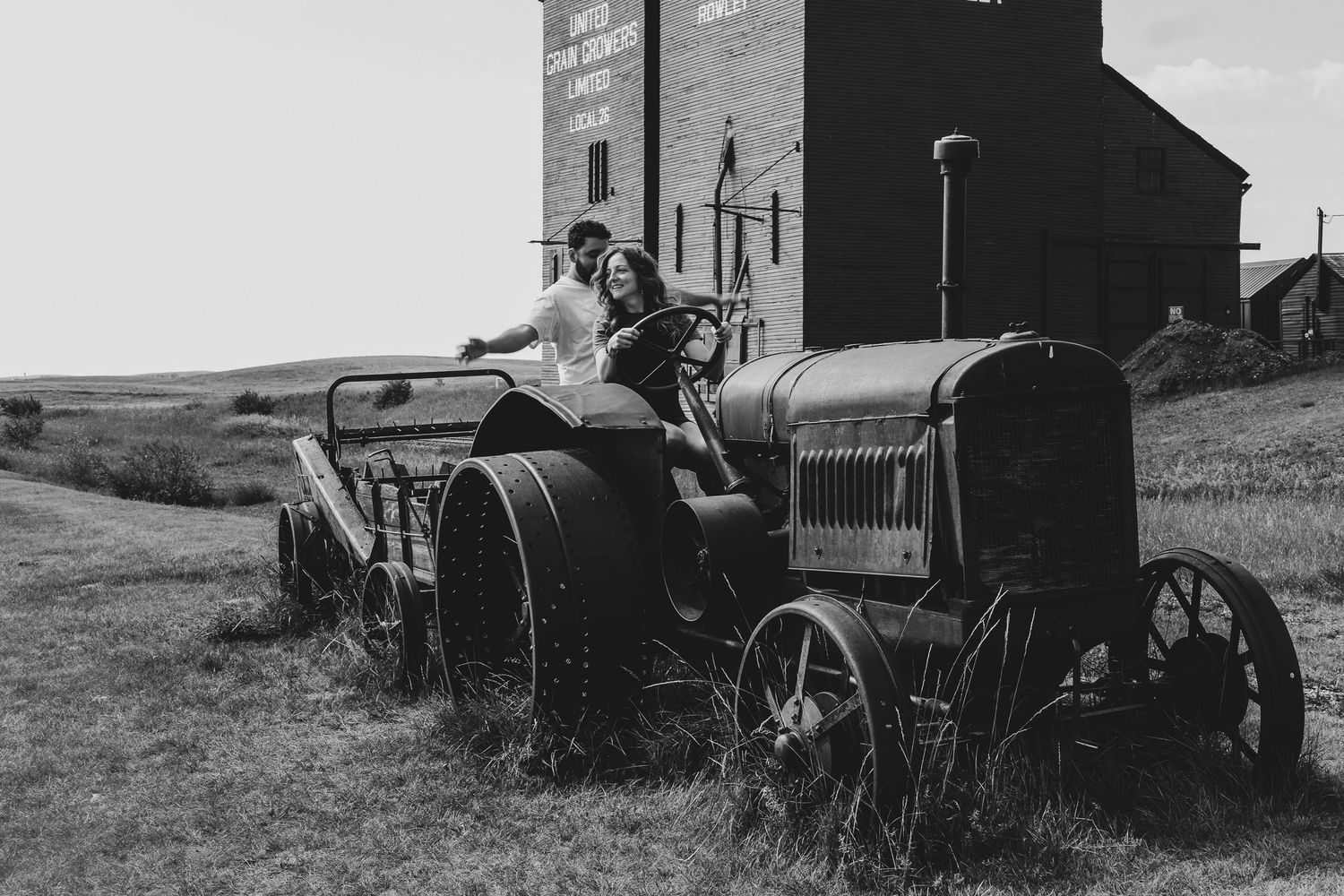 Black and white photo of a couple posing on a vintage tractor near a grain elevator in a rural setting.