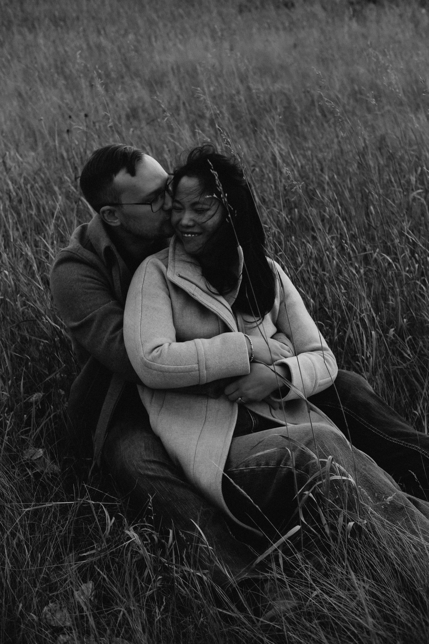 Black and white photo of a couple sitting in tall grass, man kissing womans cheek as she smiles warmly.
