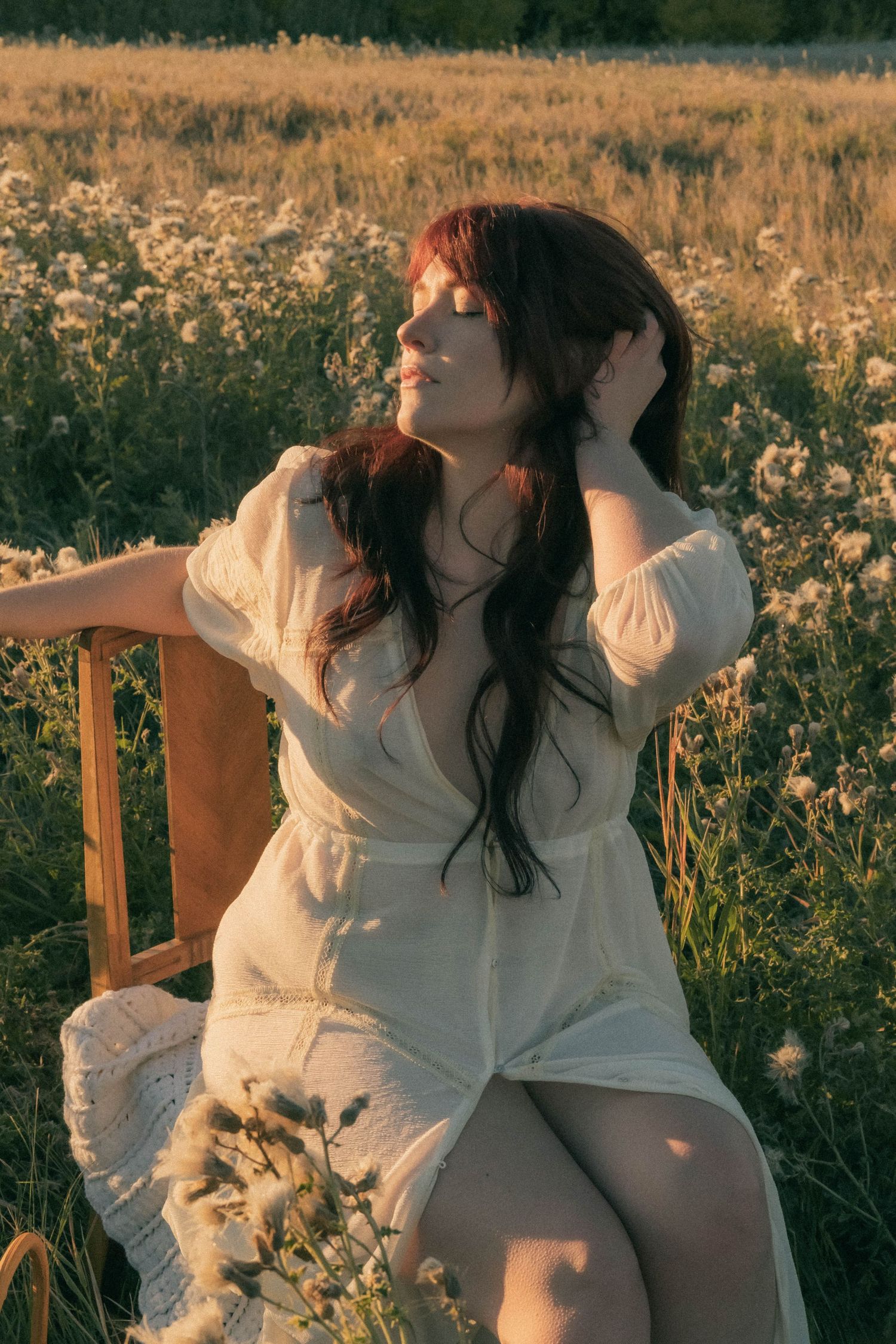 Woman in white dress sitting on chair in wildflower field at sunset.