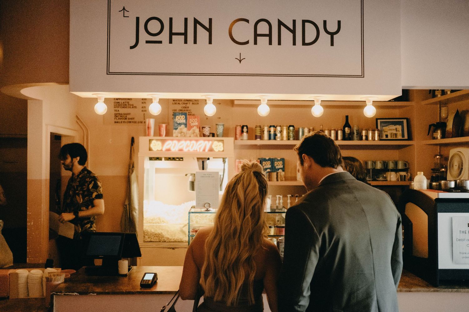 Couple ordering at John Candy cafe counter with warm Edison bulb lighting and barista.