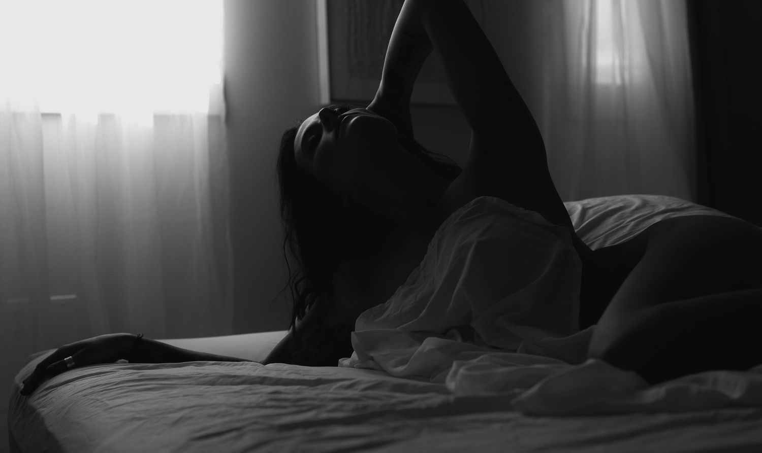 Black and white photo of a woman reclining on a bed near a window with sheer curtains.