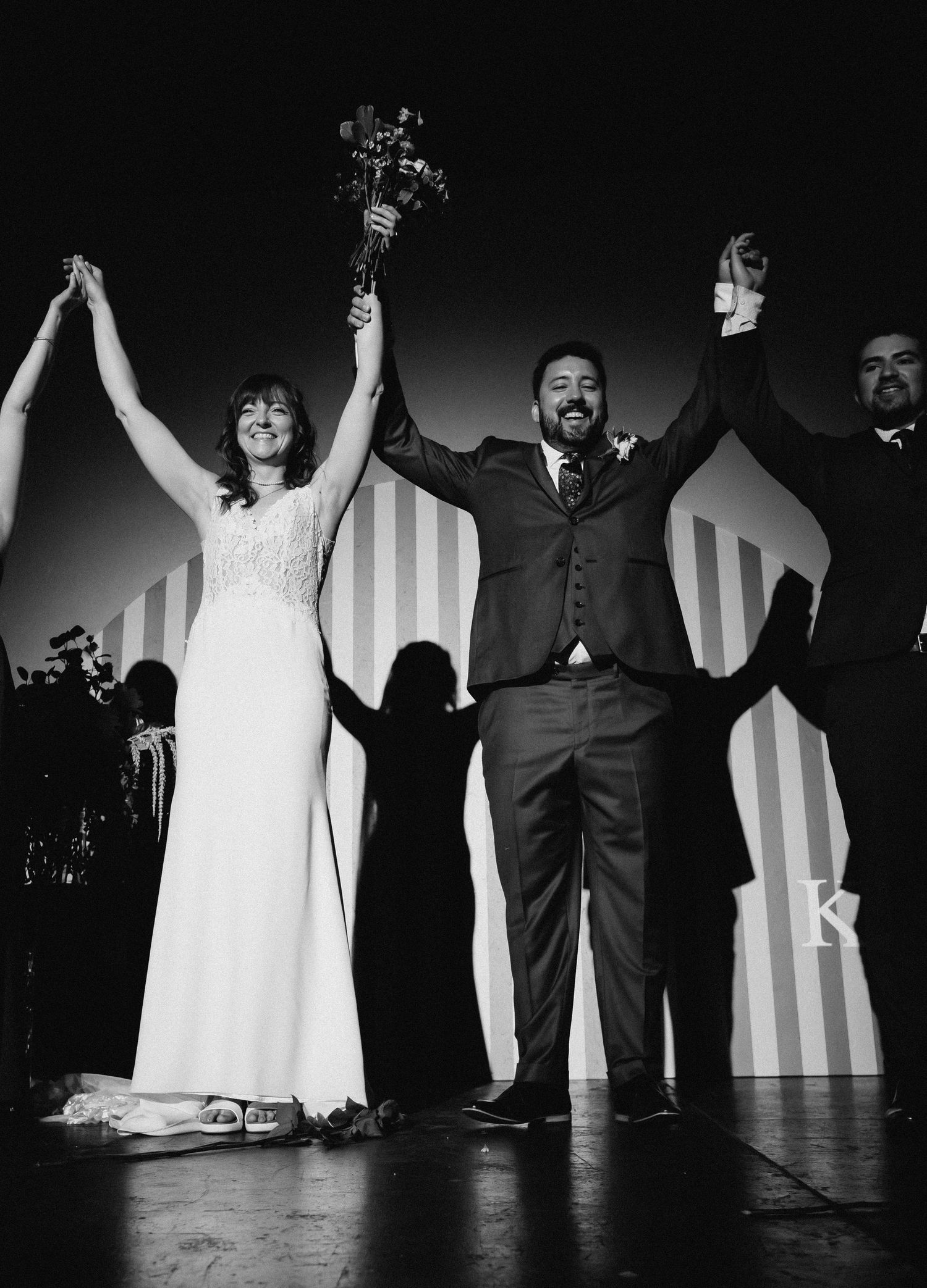 Black and white photo of a joyful bride and groom raising hands at their wedding reception on stage.
