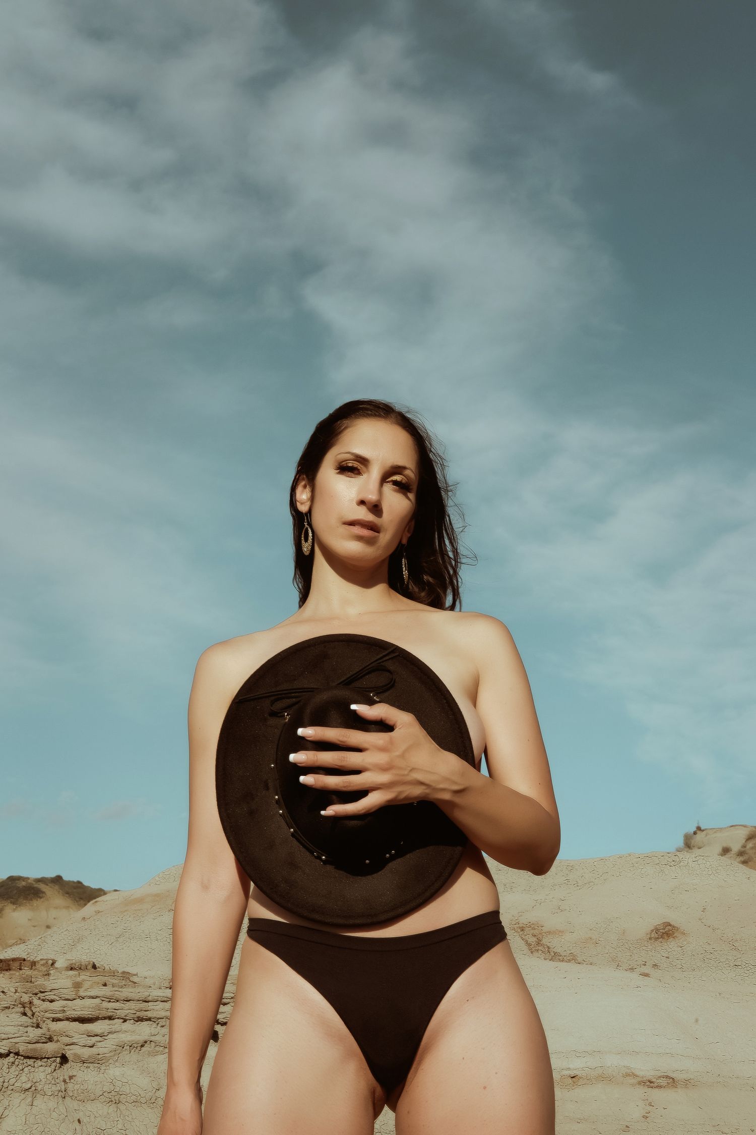 Woman in brown bikini bottom holding a hat outdoors in a sandy desert landscape under a blue cloudy sky.