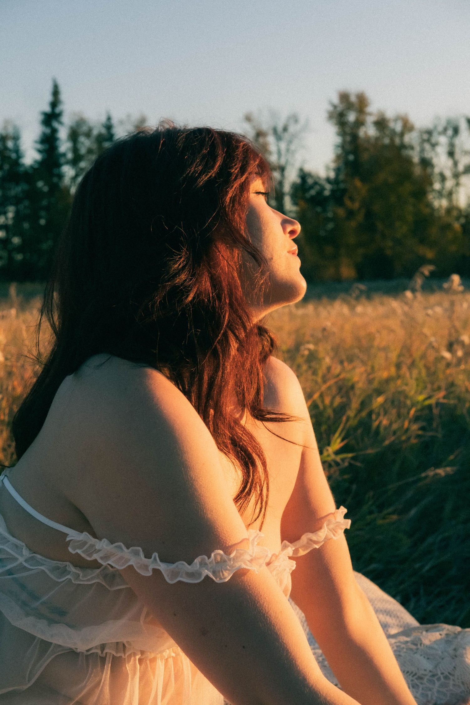 Young woman in white off-shoulder dress sitting in golden field at sunset, looking up with eyes closed.