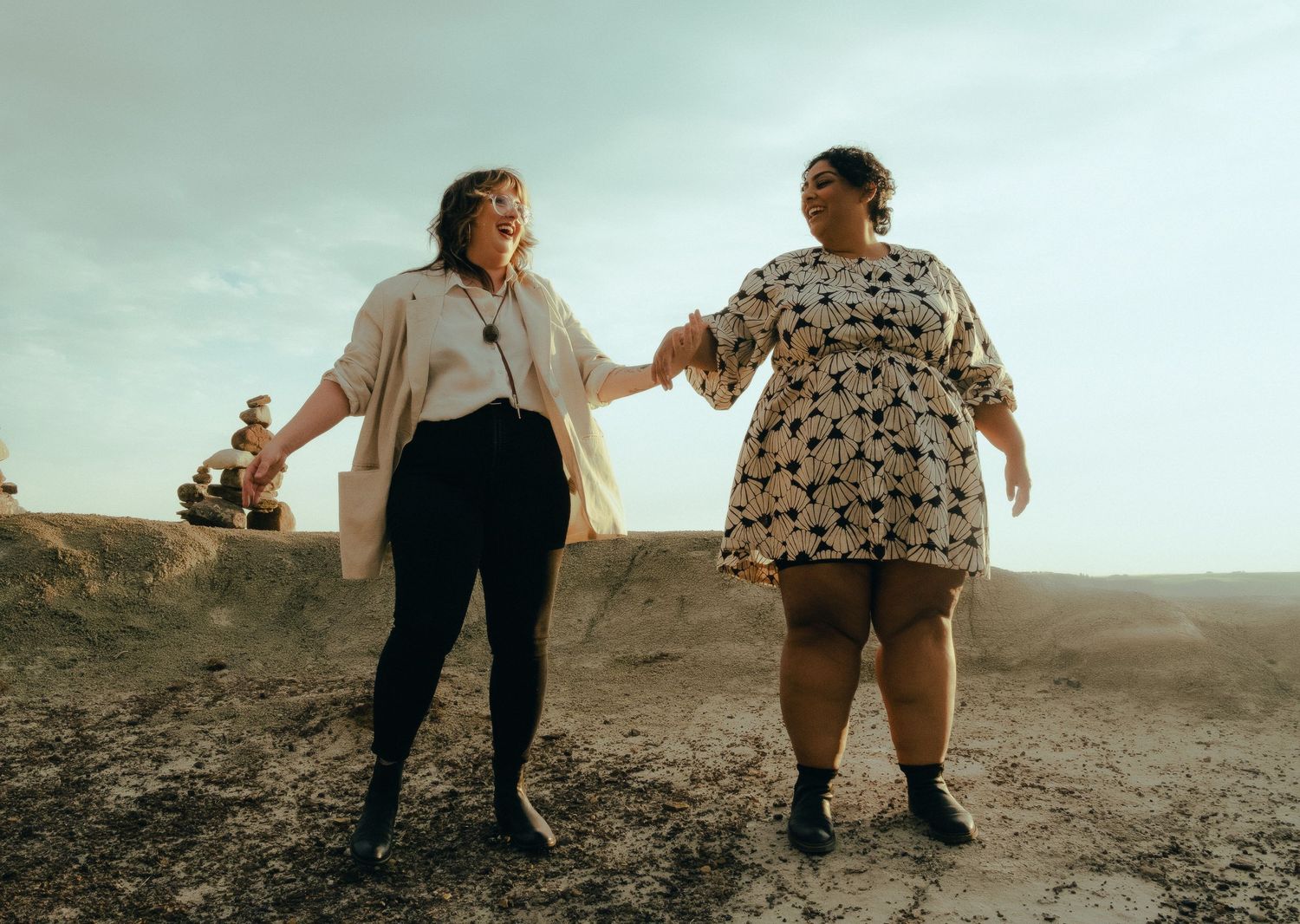 Two women holding hands and smiling joyfully on a sandy desert terrain under a dramatic sky.