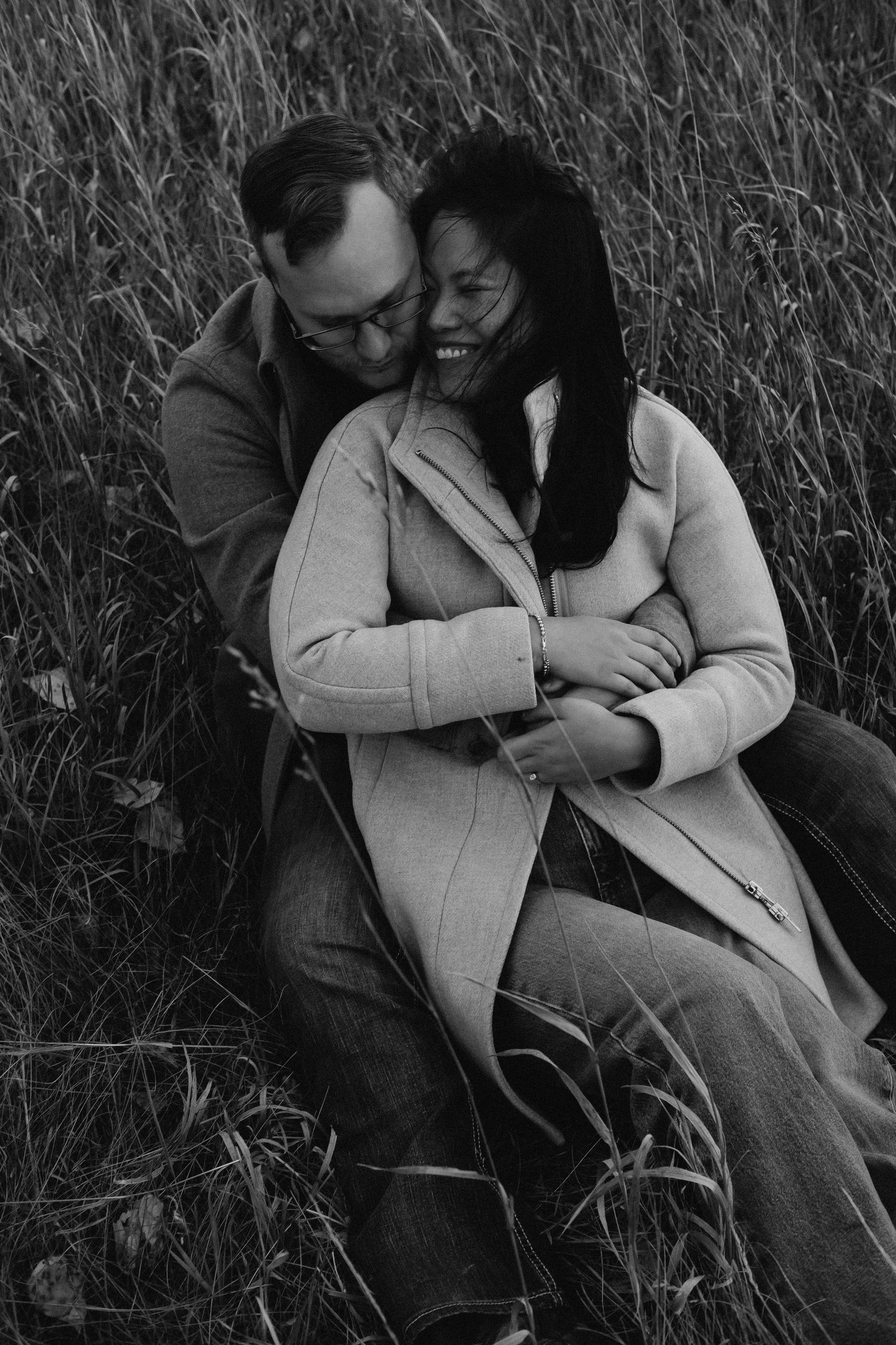 Black and white portrait of couple embracing and laughing in tall grass field.