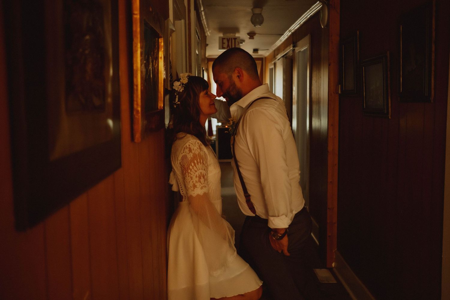 Couple sharing an intimate moment in a warmly lit vintage train corridor, bride in white lace dress, groom in suspenders.
