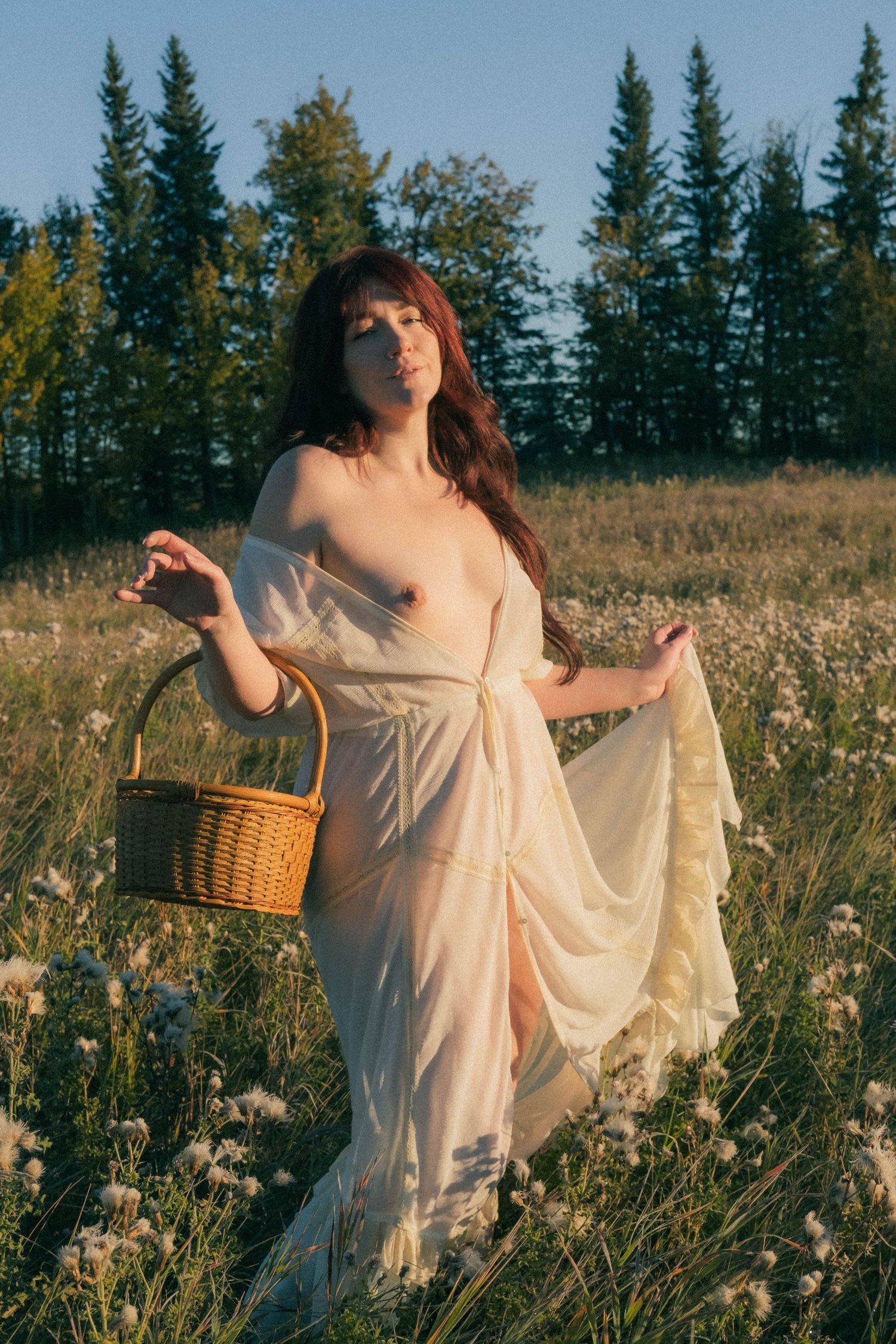 Woman in sheer white dress holding wicker basket standing in wildflower meadow at golden hour.